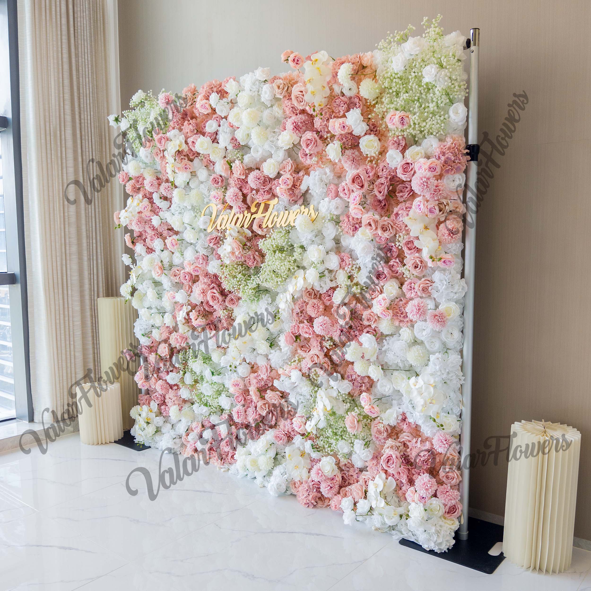 This backdrop is a rectangular floral wall supported by metal poles. It features a mix of artificial roses in soft pink, light peach, and white hues, along with clusters of baby&