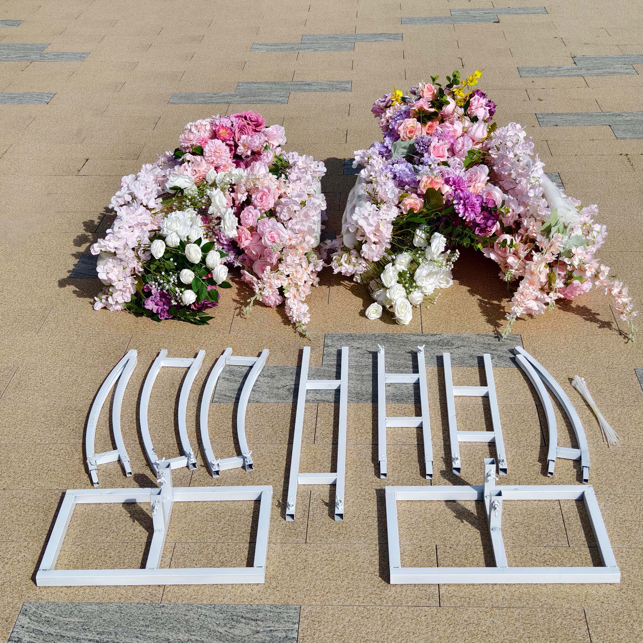 This Valar Flowers floral arch kit includes two lush, cascading arrangements of blush pink, lavender, and white blooms—including roses, hydrangeas, and wisteria—along with a white metal frame and hardware for assembly. Set on a sunlit stone surface, it creates a romantic, versatile setup ideal for weddings, engagement shoots, or elegant outdoor celebrations.