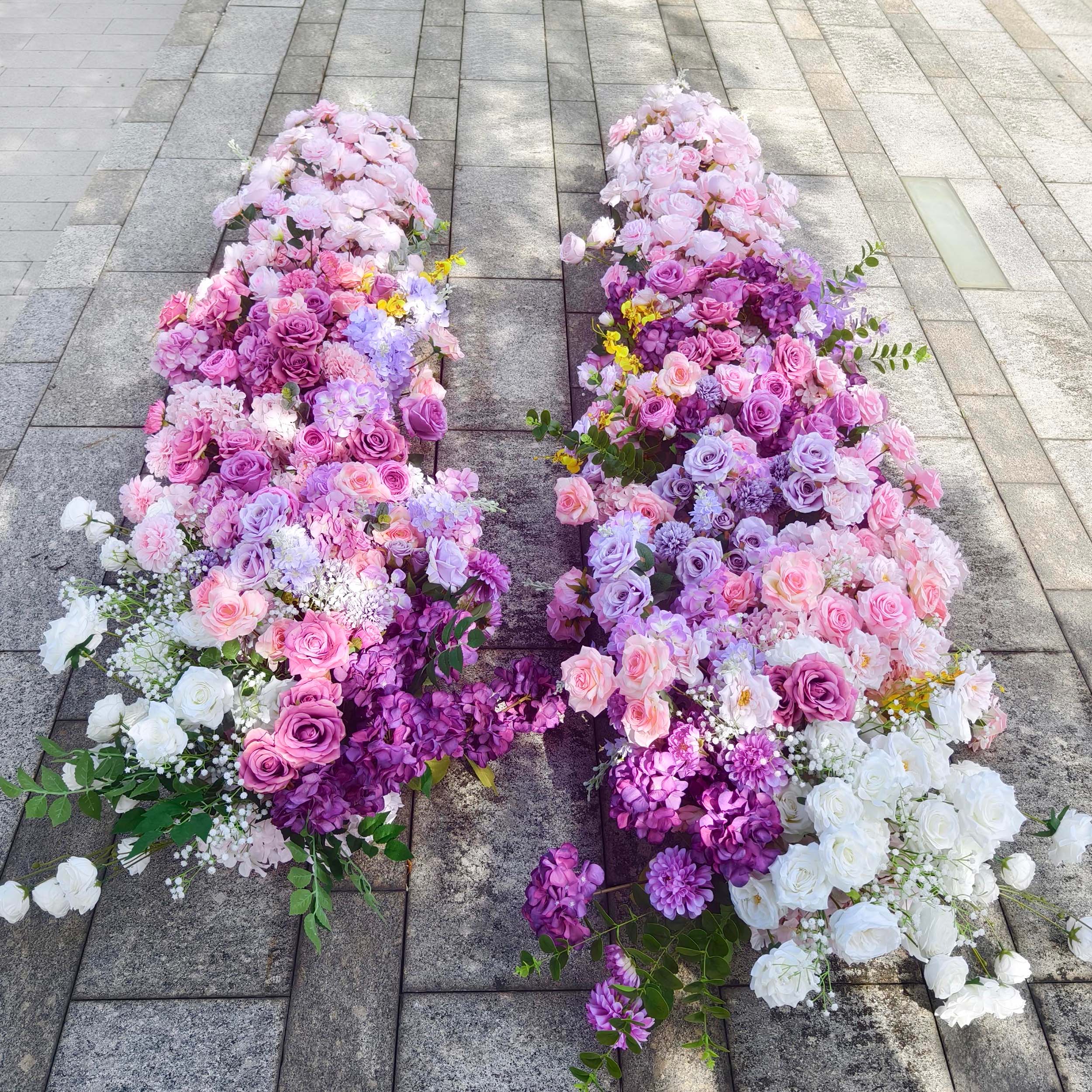 This elegant floral arrangement from Valar Flowers features a lush gradient of blush pink, lavender, and deep purple blooms—including roses, hydrangeas, and delicate sprigs—arranged in two cascading columns. Set on a sunlit stone surface, it evokes a romantic, sophisticated atmosphere ideal for weddings, engagement photoshoots, or elegant outdoor celebrations.