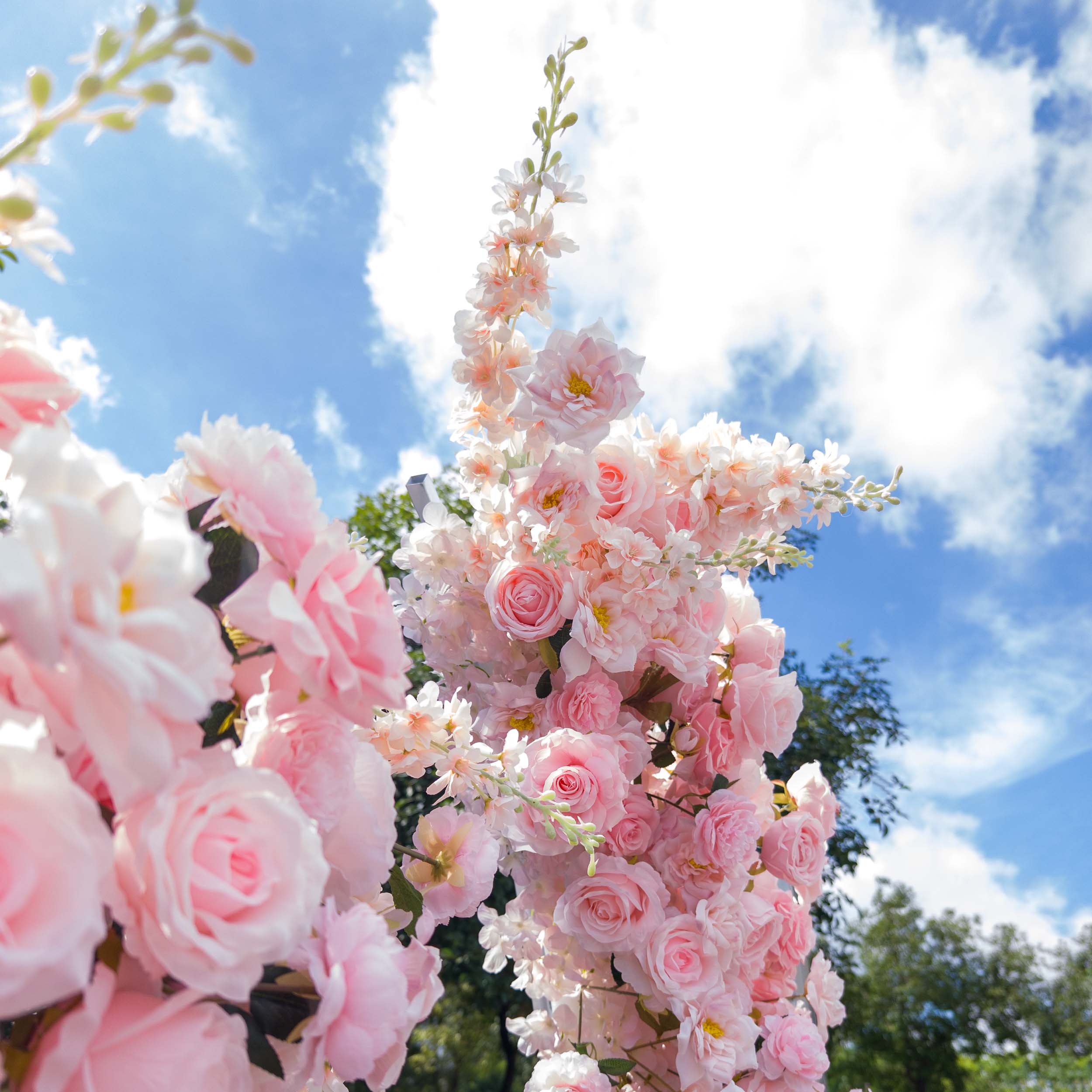 This close-up from Valar Flowers captures a lush, cascading cluster of soft pink and white blooms—including roses, peonies, and delicate sprigs—set against a bright blue sky with wispy clouds. The velvety petals and airy sprays create a romantic, dreamy atmosphere, ideal for outdoor weddings, engagement shoots, or elegant celebrations.