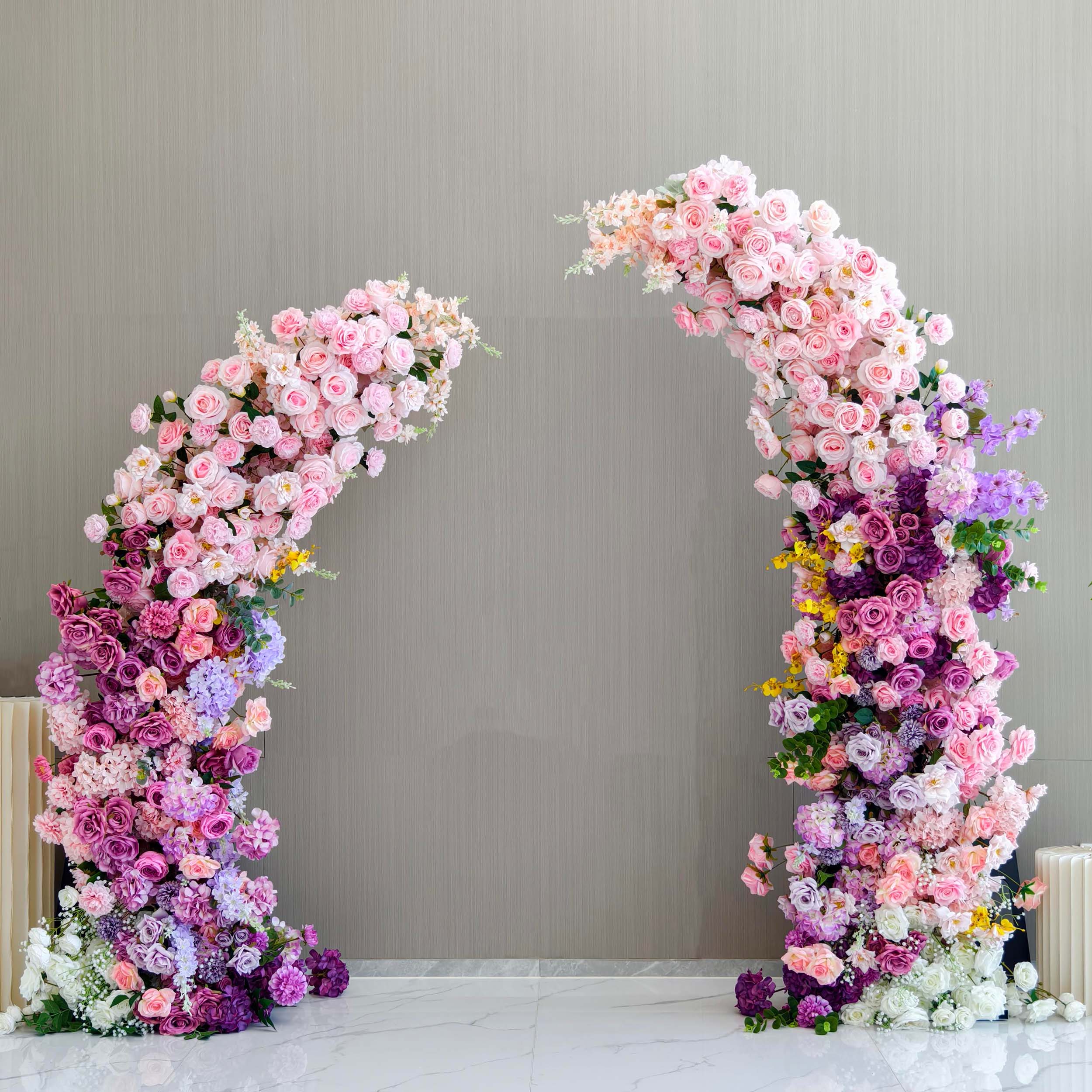 This elegant floral arch from Valar Flowers features a lush gradient of blush pink, lavender, and deep purple blooms—including roses, hydrangeas, and wisteria—arranged in two cascading columns. Set against a modern, neutral backdrop, it evokes a romantic, sophisticated atmosphere ideal for weddings, engagement photoshoots, or elegant celebrations.