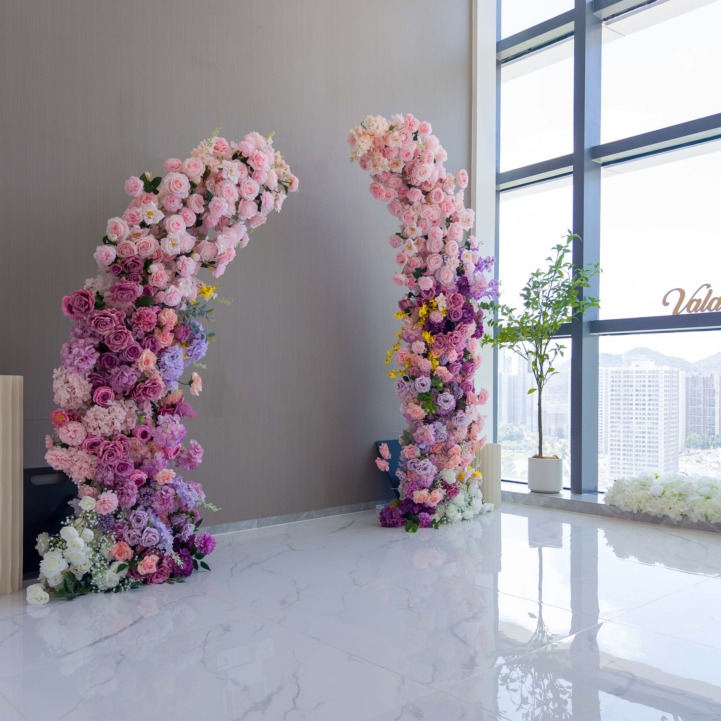 This elegant floral arch from Valar Flowers features a lush gradient of blush pink, lavender, and deep purple blooms—including roses, hydrangeas, and wisteria—arranged in two cascading columns. Set in a modern, bright interior with large windows, it evokes a romantic, sophisticated atmosphere ideal for weddings, engagement photoshoots, or elegant celebrations.