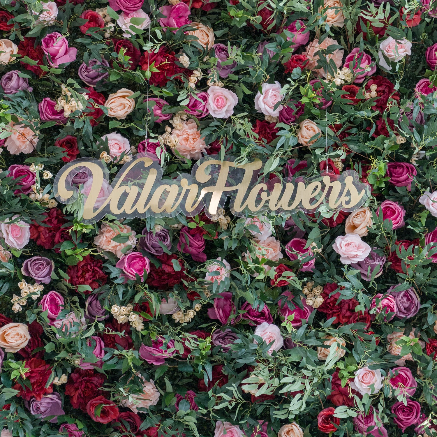 A lush artificial flower wall with pink, red, purple, and peach blooms, plus green foliage. At the center, a gold “ValarFlowers” sign on clear acrylic stands out. The vibrant, varied flowers create a romantic, decorative backdrop, perfect for events or branding displays.