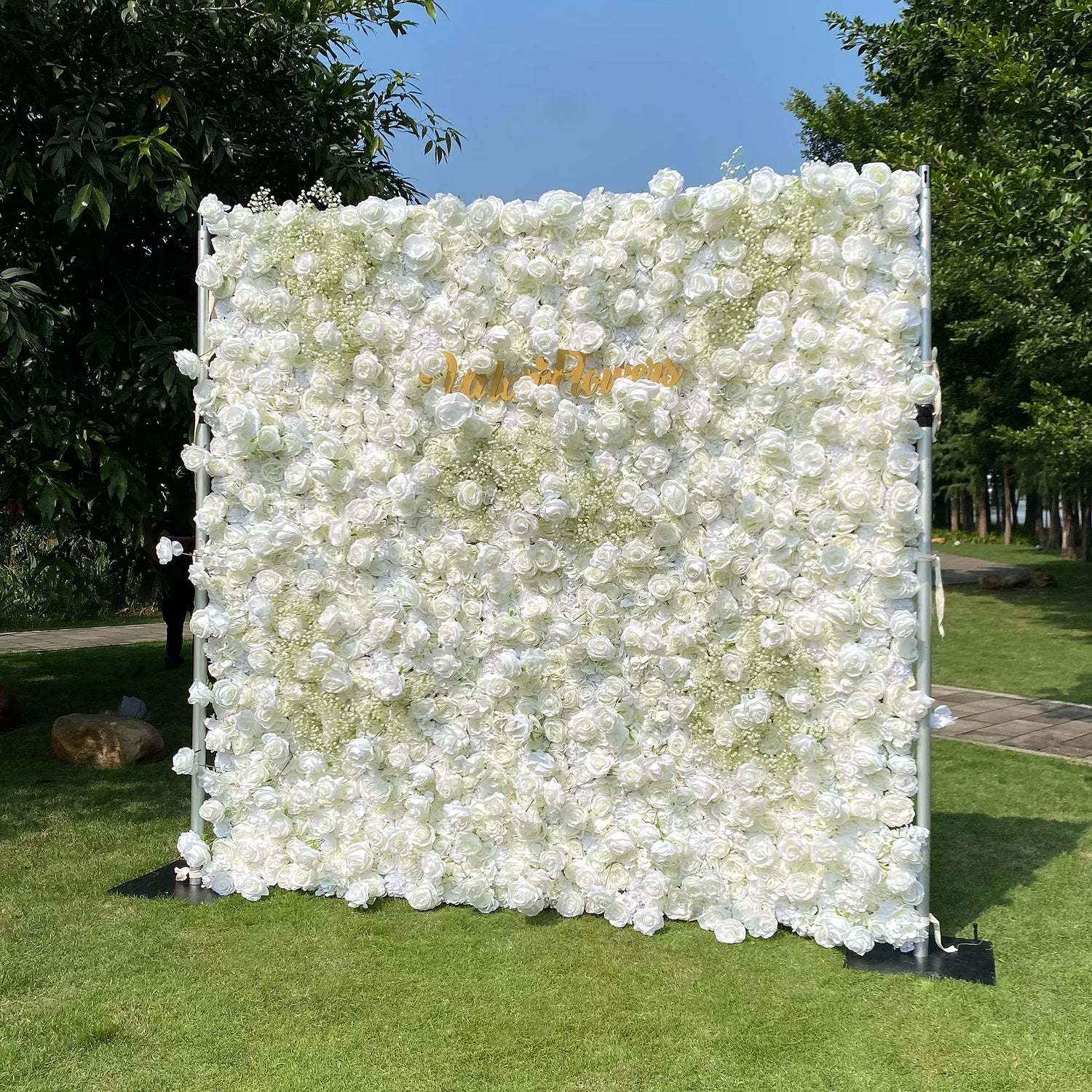 This image showcases a large floral wall densely covered with white flowers. "Valar Flowers" is displayed in golden text. Set outdoors on a lush green lawn, with trees and a clear blue sky in the background, it creates an elegant and serene atmosphere, perfect for events or decorative displays.