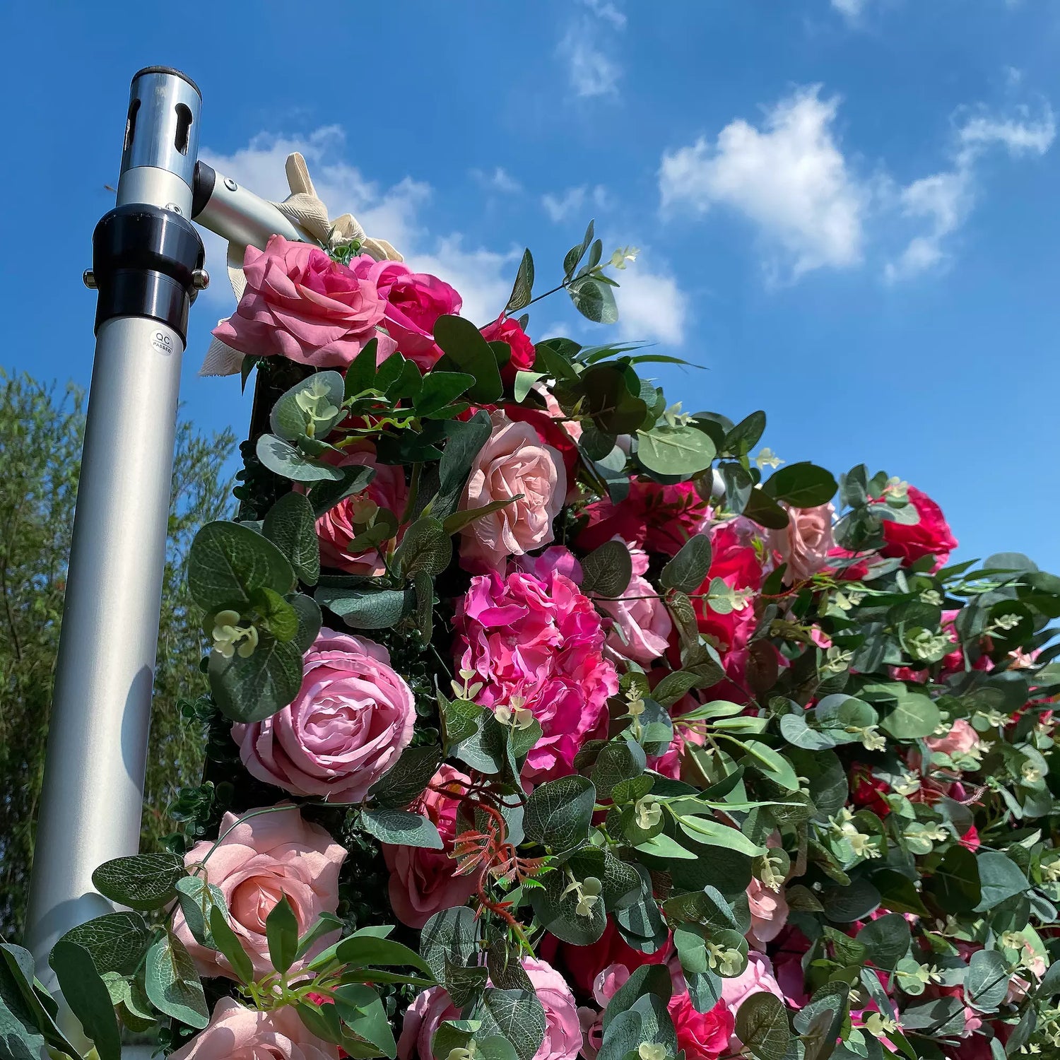 This image features a vibrant arrangement of artificial flowers, including roses in shades of pink and red, along with pink hydrangeas. Interspersed are lush green leaves, creating a dense, colorful display. The flowers are part of a structure held by a metallic pole, set against a bright blue sky with scattered clouds, exuding a cheerful and elegant vibe, perfect for decorative purposes in outdoor events.