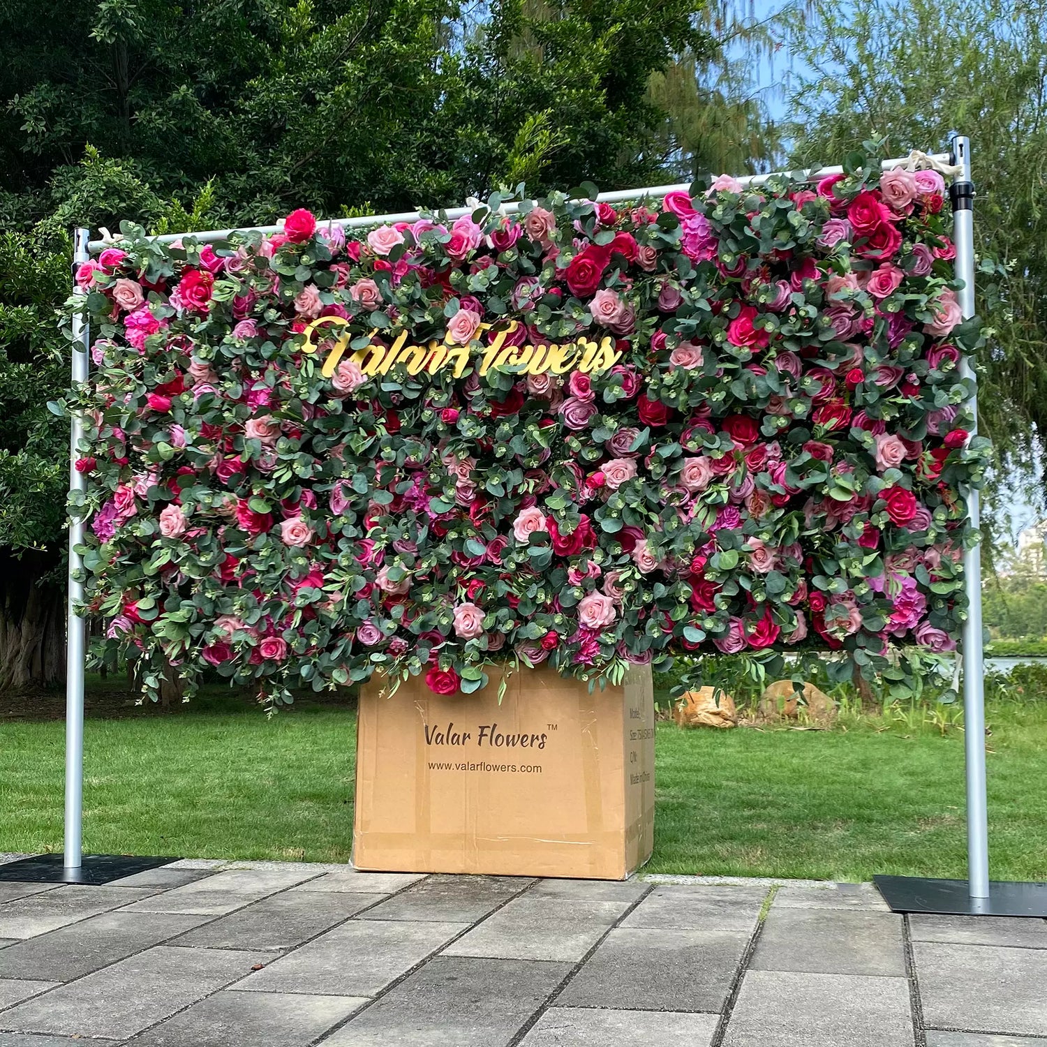 This image features a vibrant floral wall by Valar Flowers, displayed outdoors. Densely covered with roses in shades of pink, red, and purple, along with green foliage, it has a golden "Valar Flowers" sign. The backdrop includes a lawn, trees, and a glimpse of water under a bright sky, creating an elegant, nature - filled scene ideal for events.