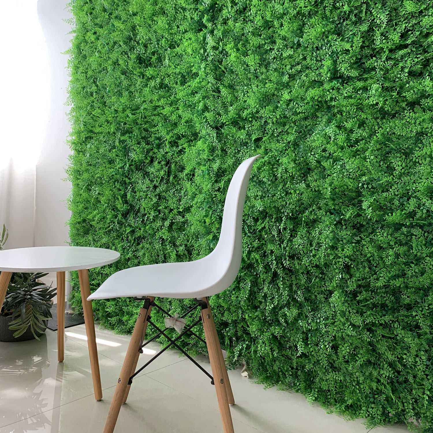 This image features an indoor scene with a lush artificial greenery wall as the focal point, showcasing dense, vibrant green foliage that creates a natural, refreshing backdrop. In the foreground, there’s a modern white chair with wooden legs and a small round white table. A potted plant adds to the organic feel, while natural light enhances the space, blending modern decor with a serene, nature-inspired atmosphere.