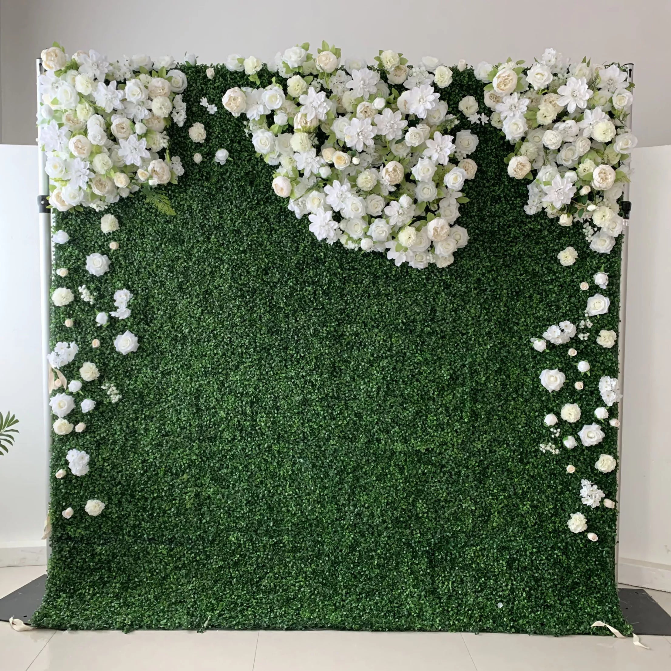 This elegant floral wall features a lush, deep green foliage base, adorned with a cascading arrangement of white and cream blooms—including roses, hydrangeas, and delicate filler flowers. The blooms form a soft, heart-shaped design at the top, with individual flowers trailing down the sides. Set against a minimalist white backdrop, the design evokes a fresh, romantic, and naturally sophisticated atmosphere ideal for weddings, photo backdrops, or elegant celebrations.