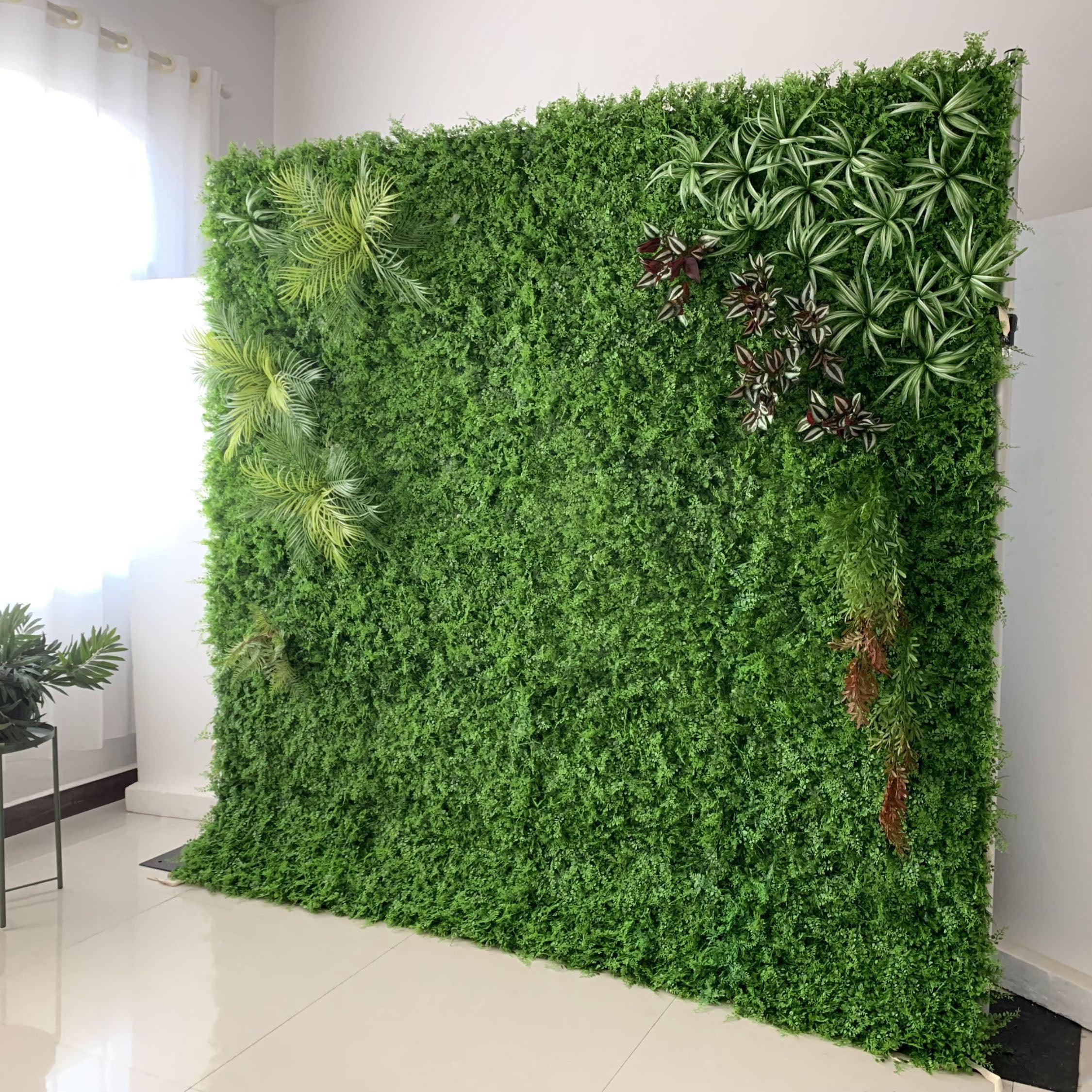 This image showcases a lush artificial greenery wall set indoors on a glossy white floor. It features a dense base of vibrant green foliage, artfully accented with varied plants like palm fronds, spiky greens, and variegated leaves. Natural light streams in from a window, enhancing the realistic textures and rich green hues. The diverse elements create a natural, refreshing display, perfect for adding organic elegance to interiors, exuding a serene and sophisticated atmosphere.