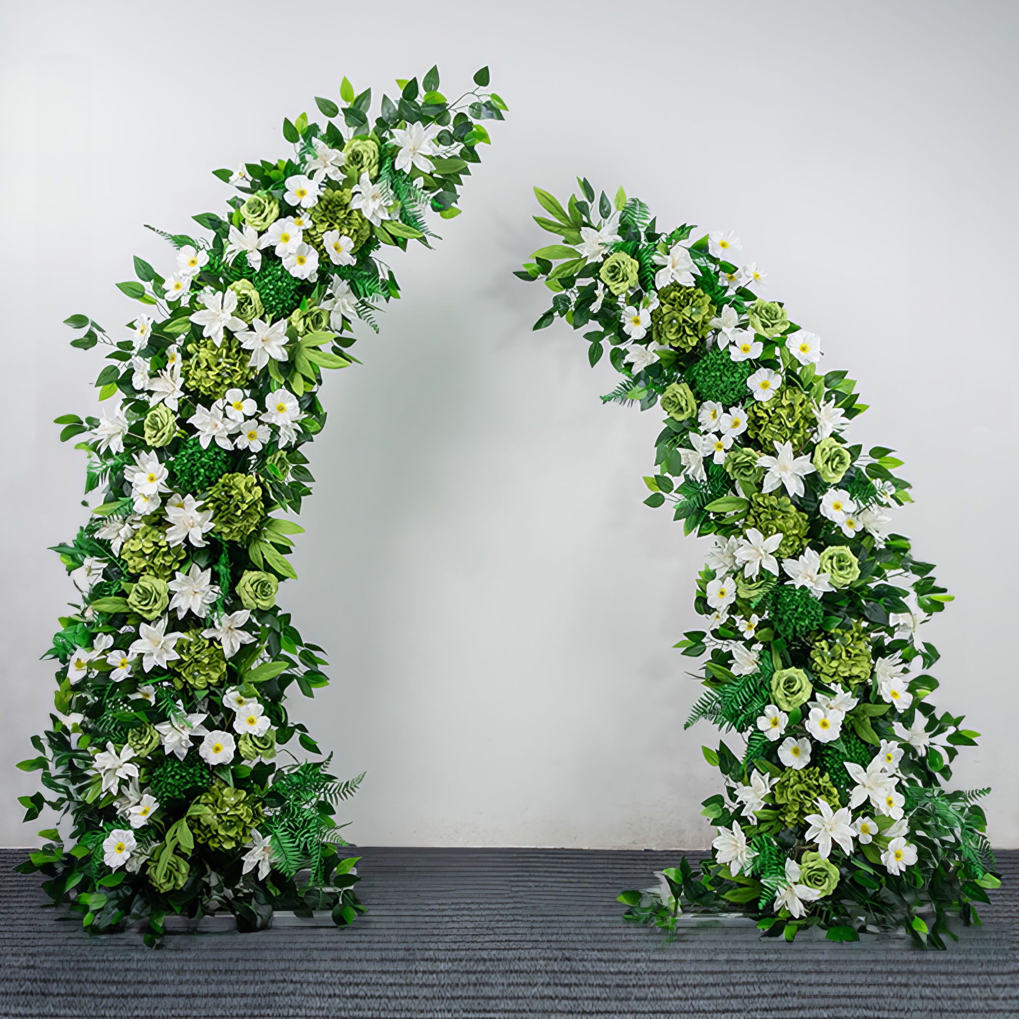 A timeless, symmetrical floral arch from Valar Flowers, crafted with a fresh palette of pure white lilies, roses, and vibrant green hydrangeas, intertwined with lush eucalyptus and fern foliage. Set against a neutral studio backdrop, it creates a serene, elegant, and eye-catching aesthetic ideal for wedding ceremonies, photo sessions, and special event decor.