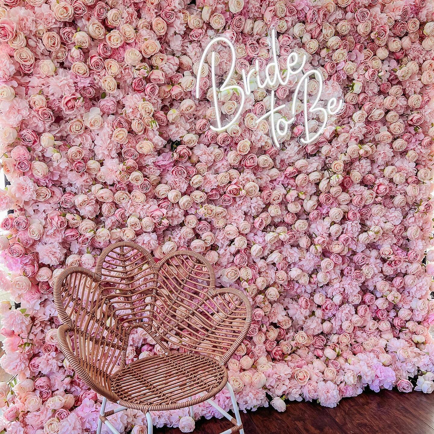 This photo showcases a stunning floral backdrop densely covered in soft pink roses. The word “Bride to Be” is prominently displayed in elegant neon lights, adding a celebratory touch. In the foreground, a heart - shaped wicker chair with a woven design sits on a dark wooden floor, complementing the romantic setting. Softly lit by natural light filtering through sheer curtains, the scene exudes charm and is perfect for bridal events.
