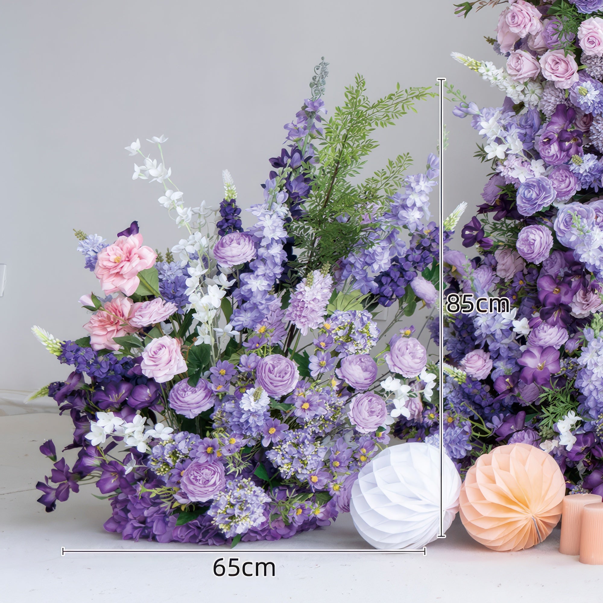 This lush floral arrangement from Valar Flowers features a rich gradient of soft pink, lavender, and deep purple blooms—including roses, hydrangeas, and delphiniums—interwoven with vibrant green foliage and delicate white accents. Standing 85cm tall and 65cm wide, it is accented by decorative paper lanterns and candles, evoking a romantic, sophisticated atmosphere ideal for weddings, engagement photoshoots, or elegant celebrations.