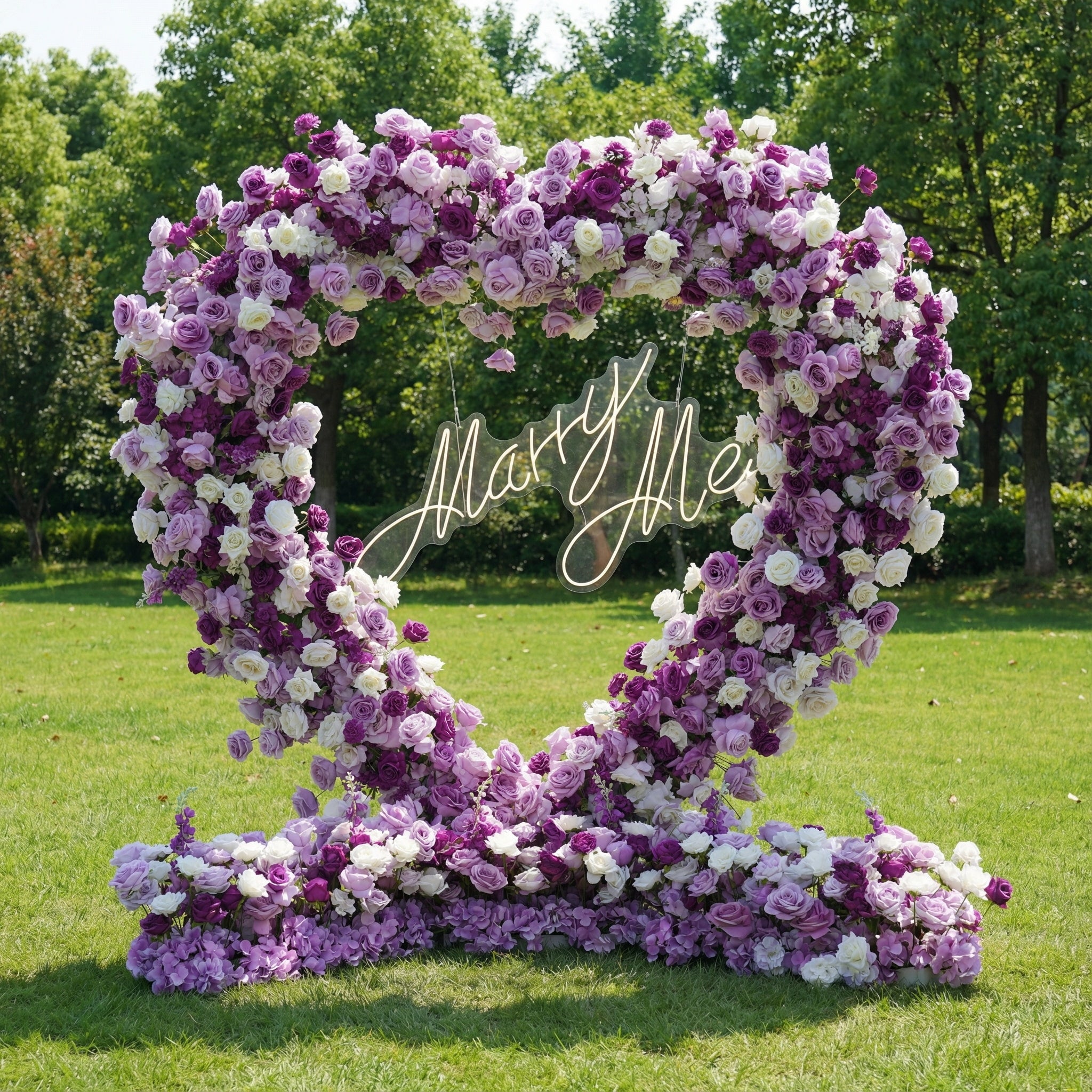 This romantic heart-shaped floral arch from **Valar Flowers** is a lush tapestry of deep purple, soft lavender, and crisp white blooms—including roses, peonies, and delicate filler flowers. Suspended at its center is a glowing "Marry Me" neon sign. Set on a sunlit green lawn framed by leafy trees, the dense, layered design evokes a dreamy, romantic atmosphere ideal for a heartfelt proposal or engagement photoshoot.