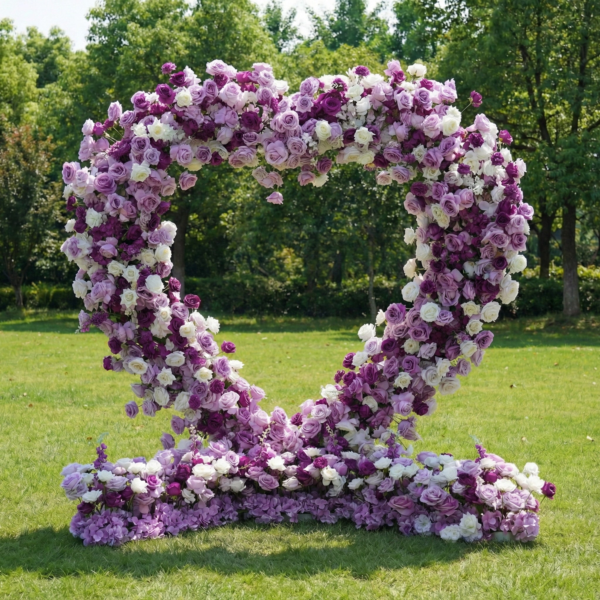 This romantic heart-shaped floral arch from **Valar Flowers** is a lush tapestry of deep purple, soft lavender, and crisp white blooms—including roses, peonies, and delicate filler flowers. Set on a sunlit green lawn framed by leafy trees, the dense, layered design creates a rich, three-dimensional texture, evoking a dreamy, romantic atmosphere ideal for a heartfelt proposal, engagement photoshoot, or elegant outdoor celebration.