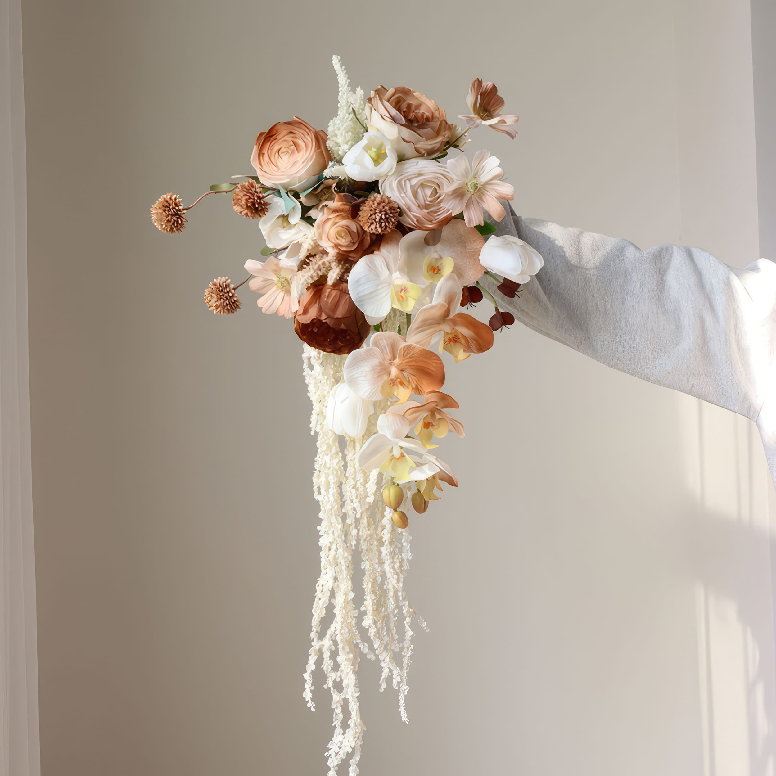 This is a cascading bridal bouquet with a warm, earthy color palette. It combines roses, orchids, daisies, and textured spherical blooms, plus trailing white amaranthus. The flowing design and neutral tones create an elegant, romantic look, perfect for weddings or special events.