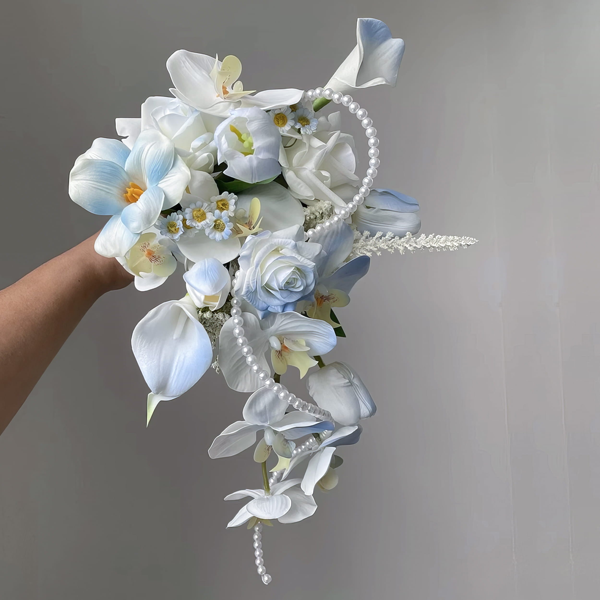 This is a cascading bridal bouquet with artificial flowers. It features white and light blue blooms like roses, calla lilies, orchids, and small daisies. Pearl strands add elegance. The flowing design and soft color palette create a romantic, dreamy look, perfect for weddings.