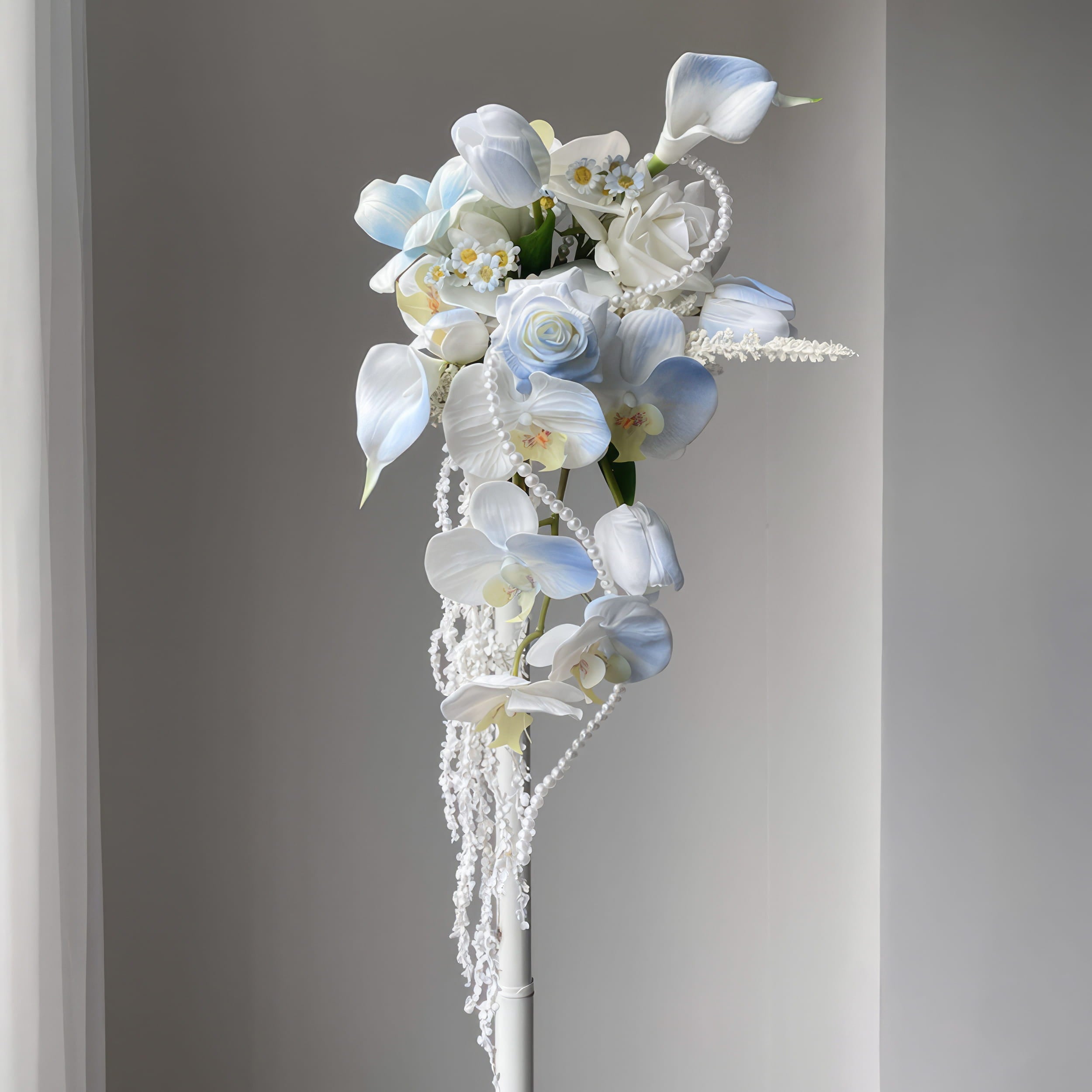 This is a cascading bridal bouquet with artificial flowers. It features white and light blue blooms like calla lilies, orchids, roses, and tiny daisies. Pearl strands and trailing white vines add elegance. The flowing design creates a romantic, dreamy look, perfect for weddings.