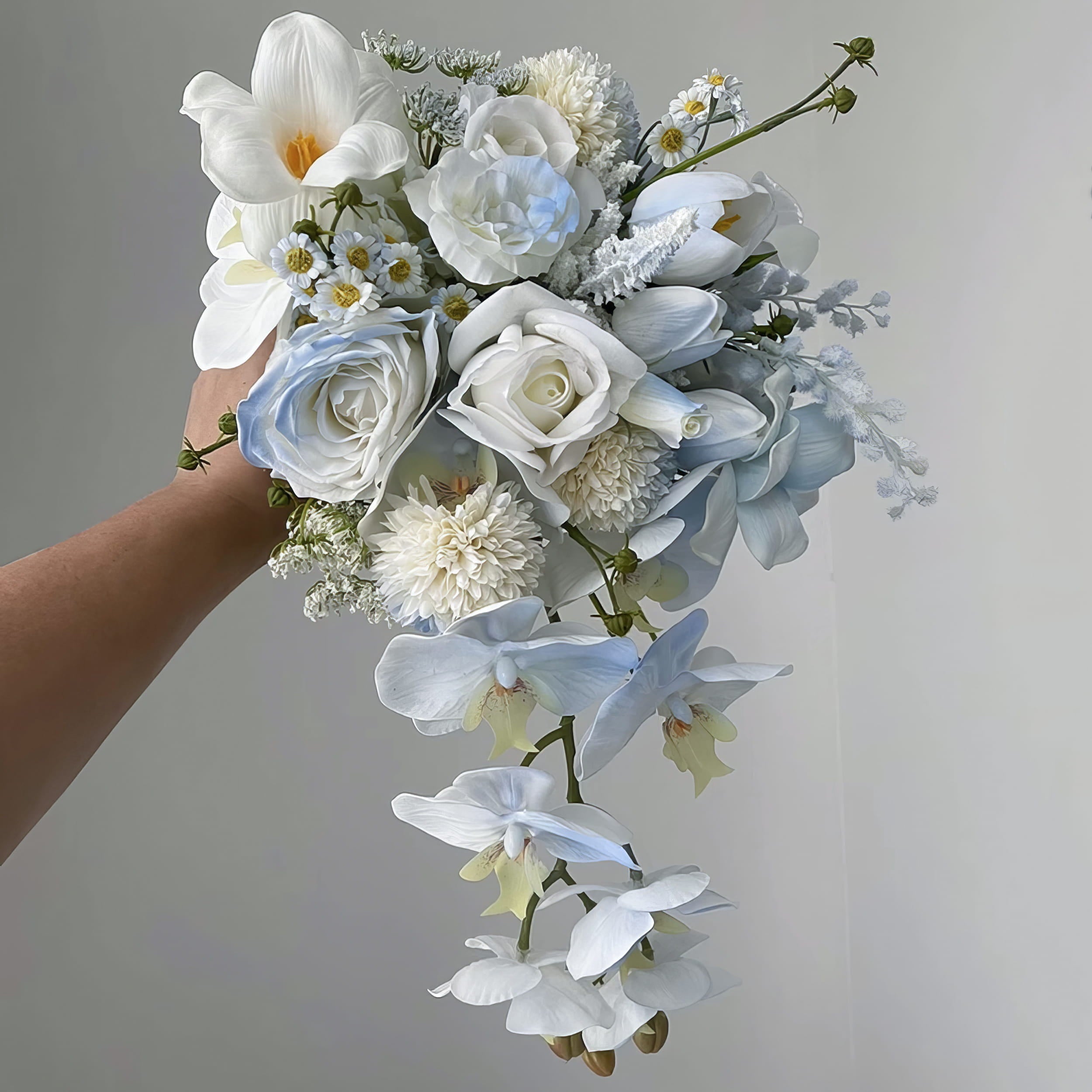 This is a cascading bridal bouquet. It combines white lilies, light - blue roses, delicate daisies, fluffy chrysanthemums, and trailing blue orchids. The soft color palette and flowing design create an elegant, romantic look, perfect for weddings.