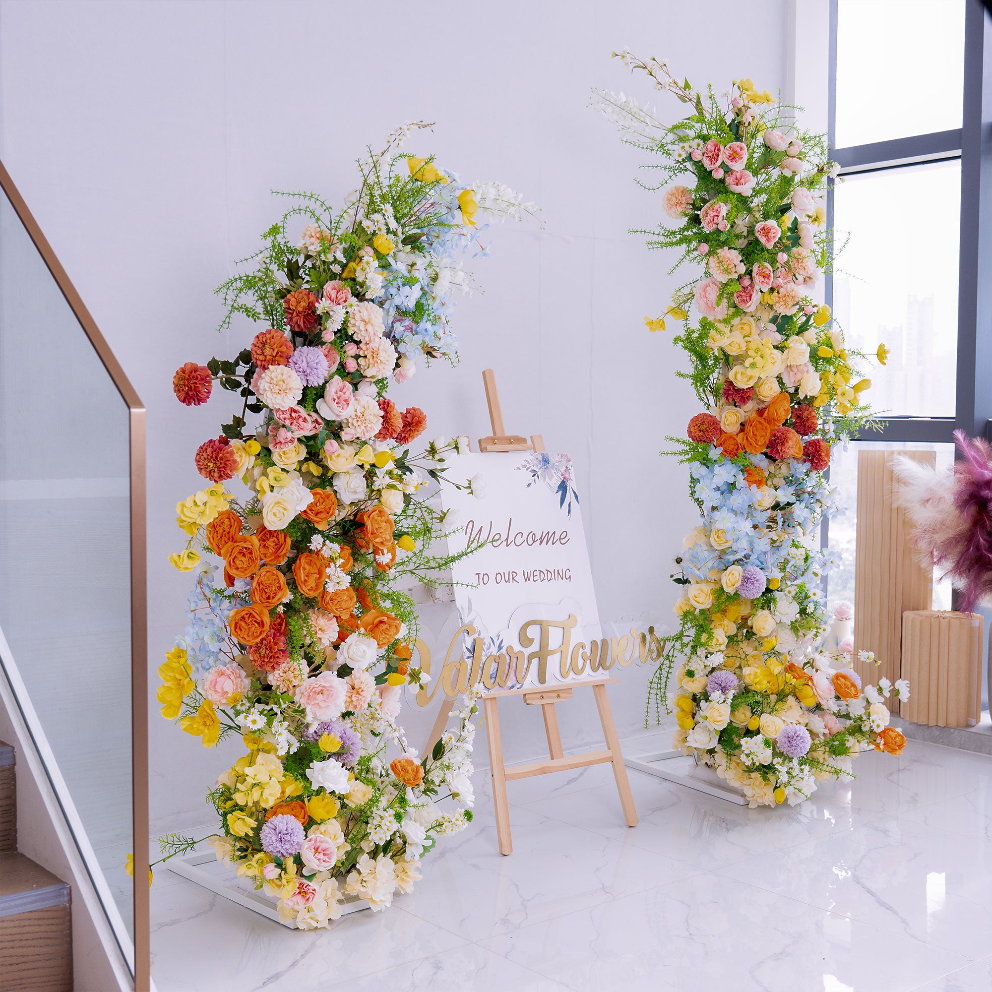 This wedding - themed setup features two large floral installations flanking a “Welcome to Our Wedding” sign. Flowers include roses, peonies, dahlias, and delphiniums, in vibrant hues like orange, yellow, pink, and blue. The lush, colorful blooms create a cheerful, romantic vibe, perfect for greeting guests and setting an elegant, festive tone for the celebration.