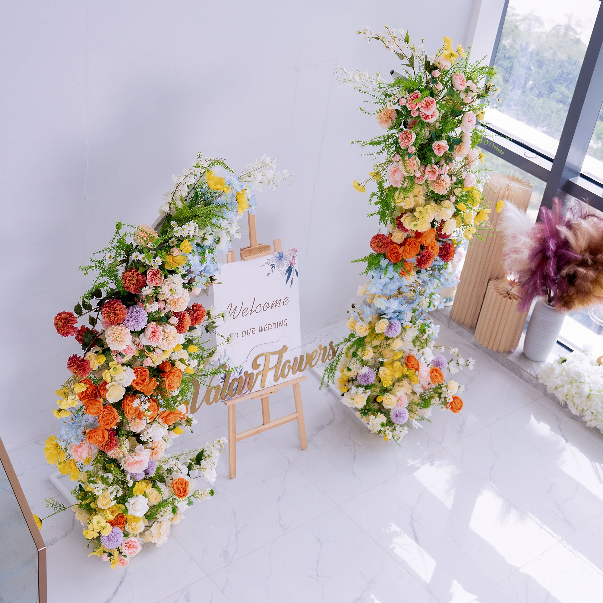 This wedding - themed setup features two large floral installations flanking a “Welcome to Our Wedding” sign. Flowers include roses, peonies, chrysanthemums, and delphinium, in vibrant hues like orange, yellow, pink, and blue. The lush, colorful blooms create a cheerful, romantic vibe, perfect for greeting guests and setting an elegant, festive tone for the celebration.