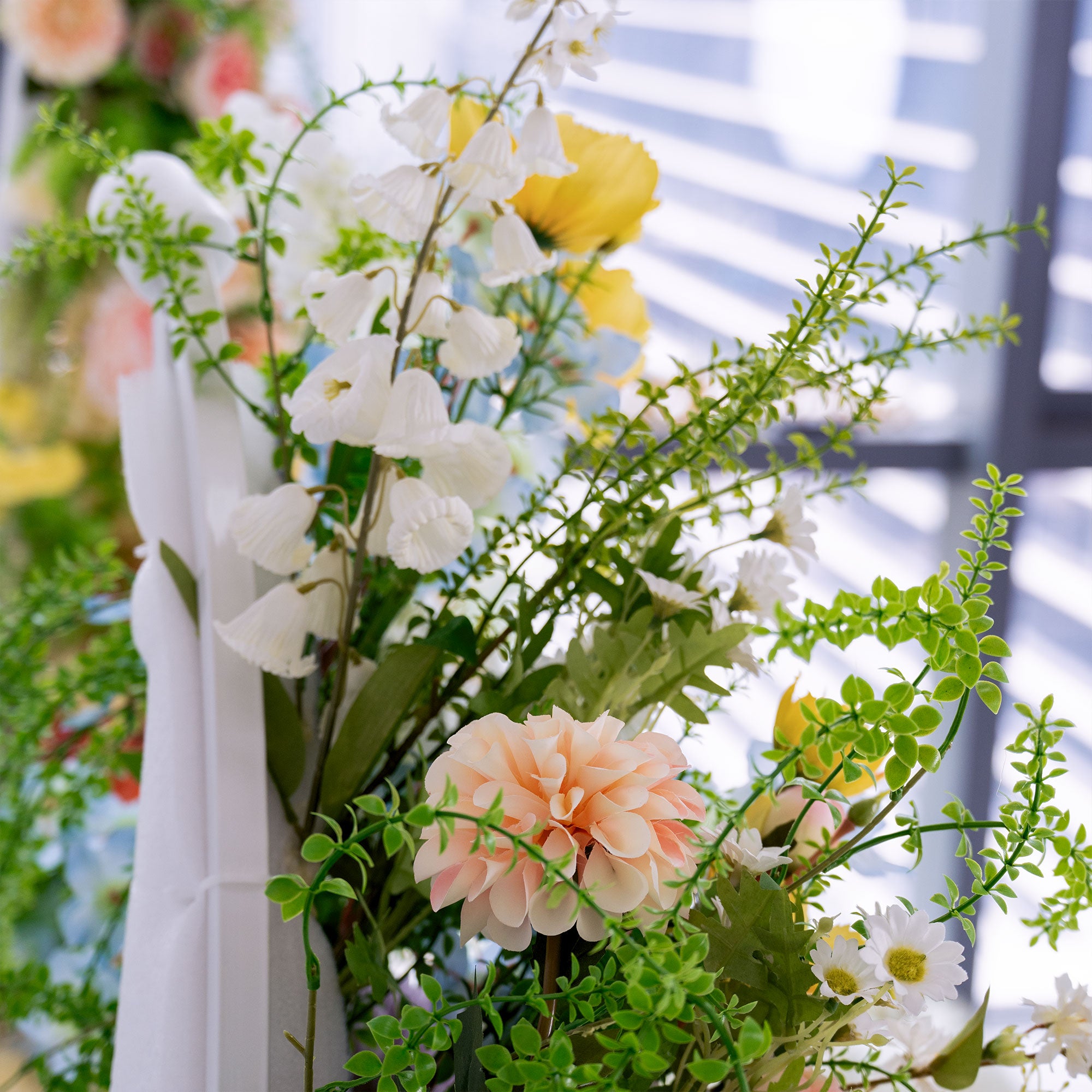 This photo showcases a beautiful arrangement of artificial flowers. It includes delicate white bell - shaped blooms, soft peach - colored dahlias, sunny yellow flowers, and small white daisies, all complemented by lush green foliage. The pastel and vibrant hues blend to create a fresh, cheerful vibe, perfect for adding a touch of natural beauty to events like weddings, parties, or home decor. The realistic textures of the faux flowers offer a long - lasting, elegant decorative option.