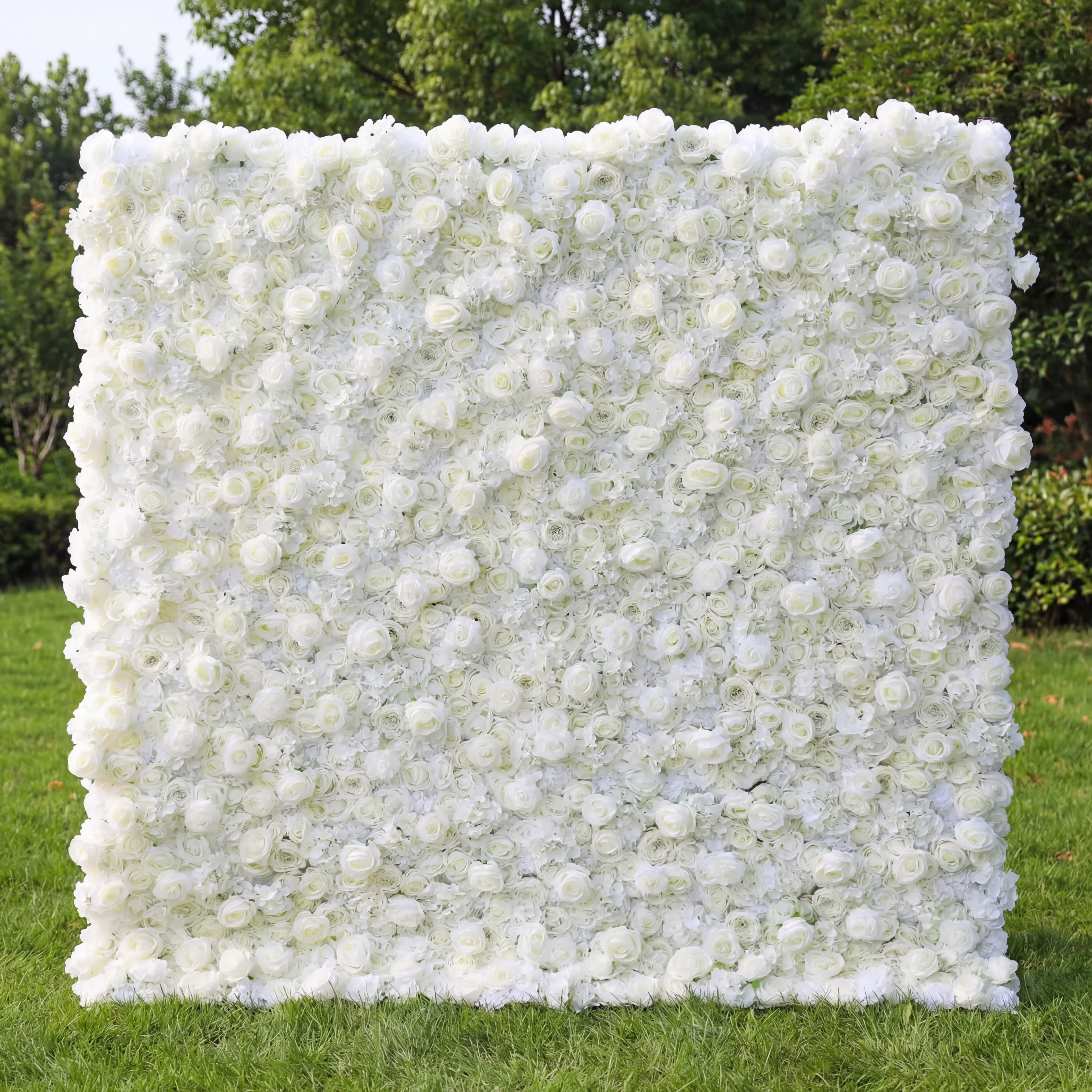 This image showcases a stunning white flower wall by Valar Flowers, composed of dense artificial white roses and blooms. Set outdoors on lush green grass with trees in the background, it exudes elegance and is ideal for events like weddings. The meticulous arrangement highlights Valar Flowers’ craftsmanship in creating sophisticated, eye-catching floral decor.