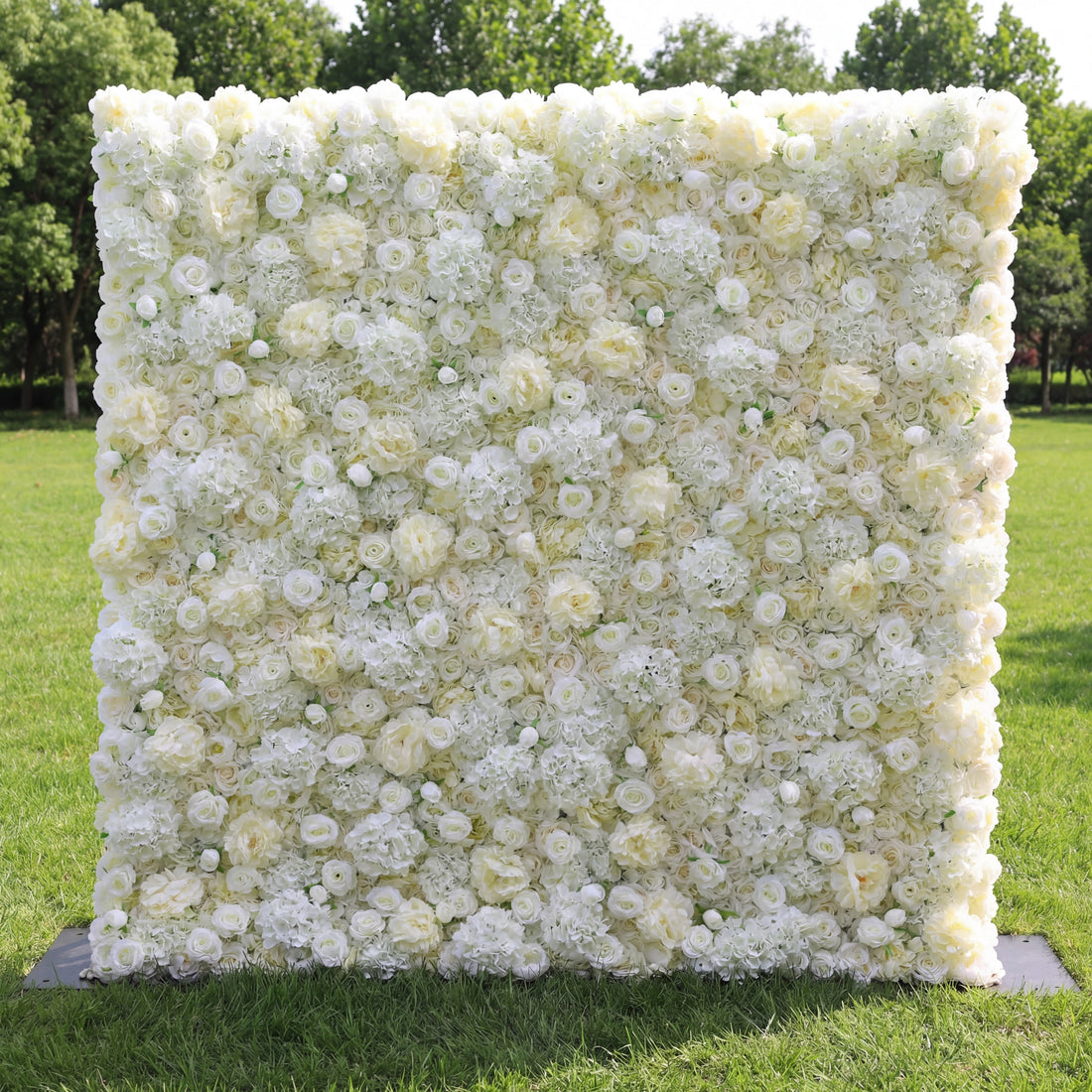 This elegant outdoor floral backdrop features a dense, velvety arrangement of artificial white and cream roses, layered with delicate filler blooms to create a lush, three-dimensional texture. Set on a sunlit green lawn and framed by leafy trees, its soft, luminous palette exudes timeless sophistication and romance, making it a stunning focal point for weddings, engagement photos, or upscale events.