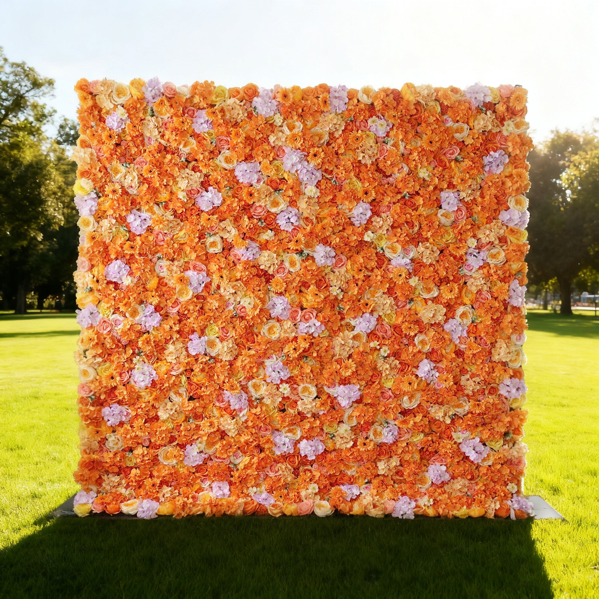 This image showcases a vibrant floral wall by Valar Flowers, set outdoors on a lush green lawn. Densely packed with artificial flowers in warm hues of orange, along with accents of pink and other complementary shades, it creates a lively and eye-catching display. Bathed in sunlight, with trees and a bright sky in the background, the floral wall exudes a cheerful and elegant atmosphere, perfect for decorative use in events like weddings or photo shoots.