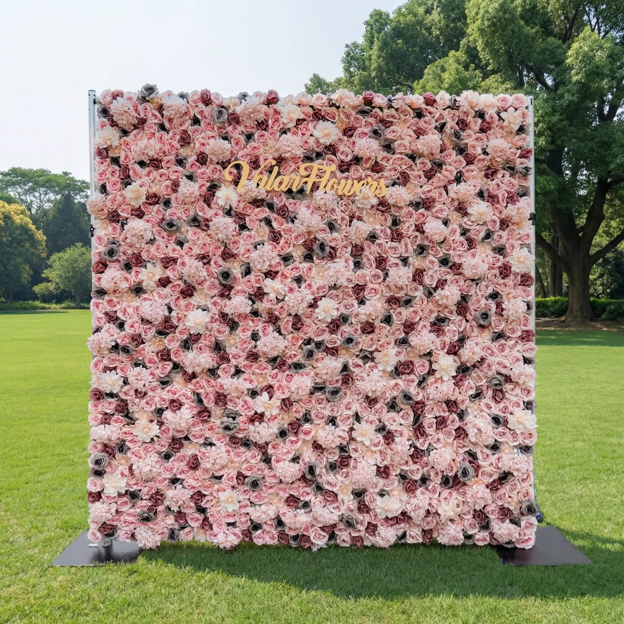 This Valar Flowers floral wall is a lush, romantic tapestry of blush pink, creamy white, and deep rose blooms, set on a sturdy metal frame atop green grass. Adorned with a gold script sign, it creates a soft, elegant backdrop ideal for outdoor weddings, photo booths, and romantic event decor.