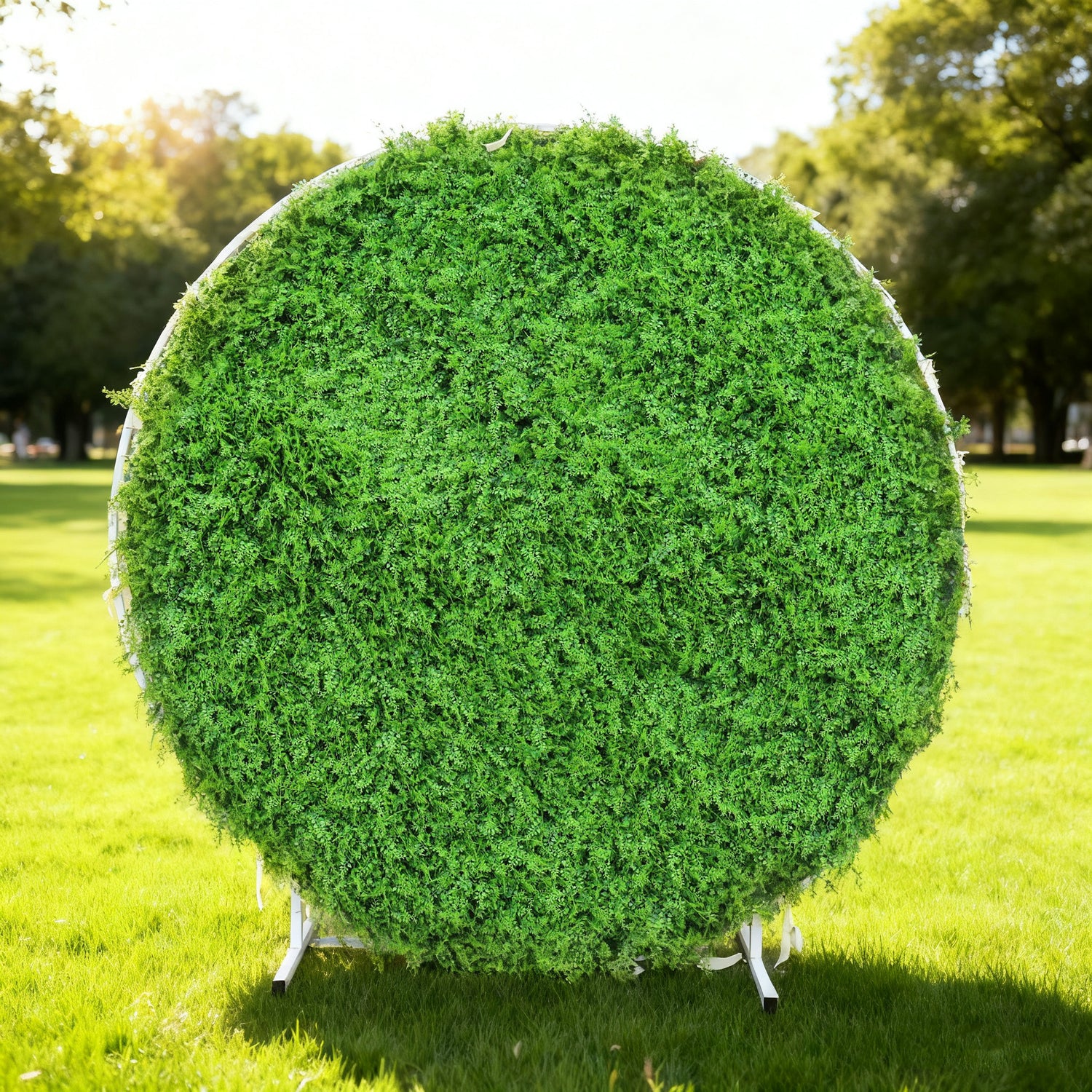 This image showcases a round artificial greenery backdrop by Valar Flowers, set outdoors on a lush green lawn. It features dense, textured green foliage, creating a natural and vibrant display. Bathed in sunlight, this circular piece is perfect for adding a touch of organic elegance to events or photo setups, exuding a serene and sophisticated atmosphere.