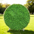 This image showcases a round artificial greenery backdrop by Valar Flowers, set outdoors on a lush green lawn. It features dense, textured green foliage, creating a natural and vibrant display. Bathed in sunlight, this circular piece is perfect for adding a touch of organic elegance to events or photo setups, exuding a serene and sophisticated atmosphere.