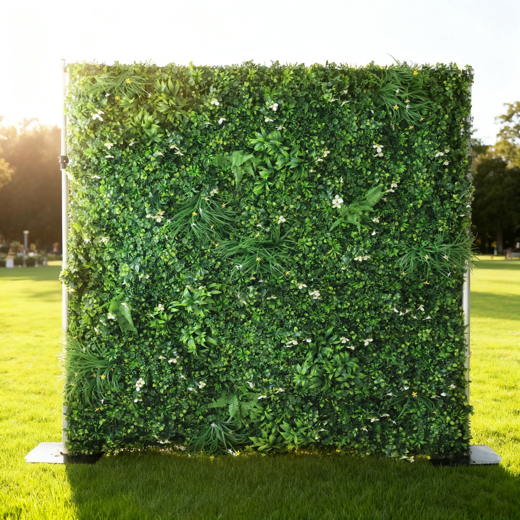 This image showcases a lush artificial greenery wall by ValarFlowers, set outdoors on a vibrant lawn. It features a dense arrangement of varied green foliage, interspersed with delicate white blooms and ferns, creating a rich, natural, and visually captivating display. Bathed in sunlight, this piece is perfect for adding organic elegance to events or spaces, exuding a serene and sophisticated atmosphere.