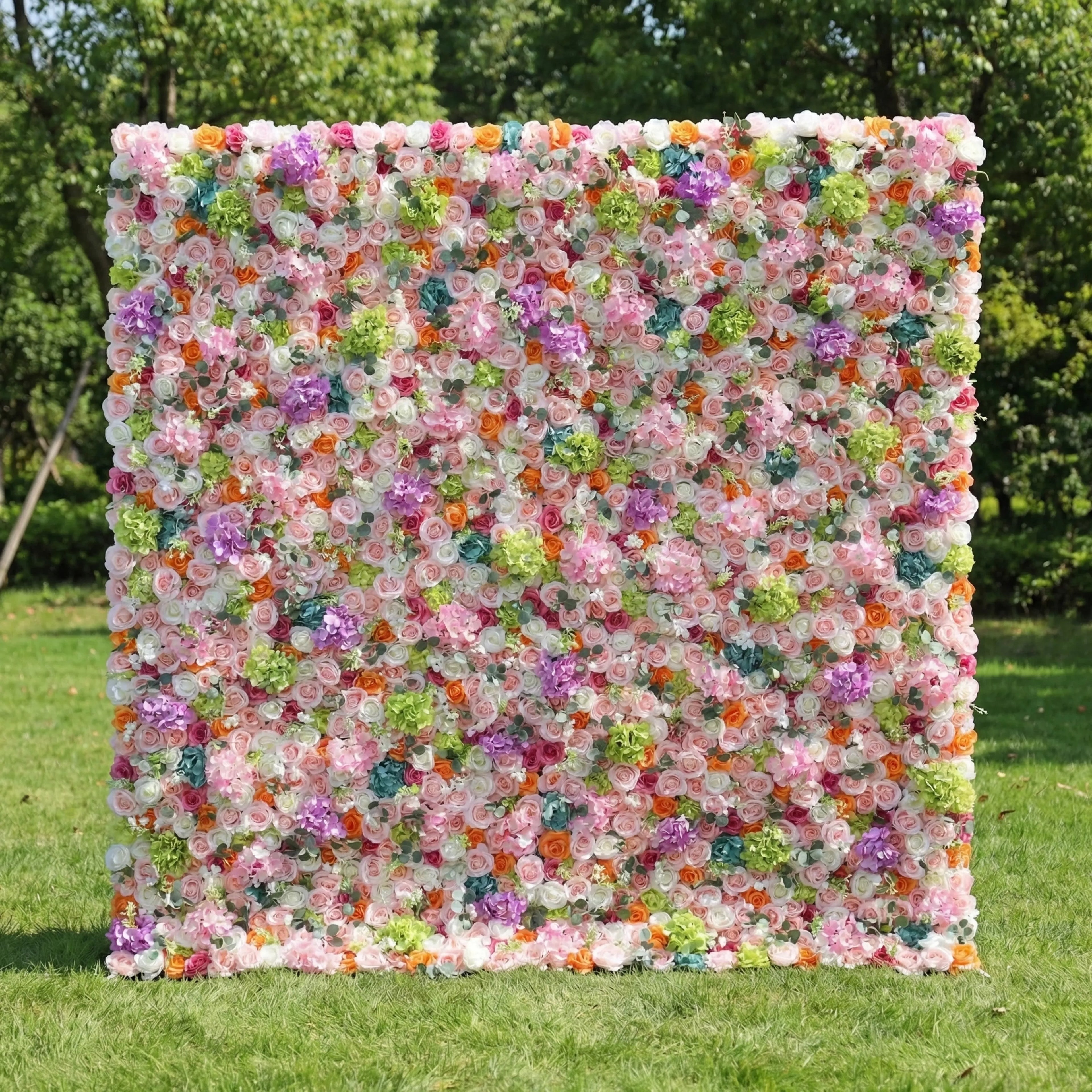 This is a vibrant, multi-colored artificial flower wall backdrop from **Valar Flowers**, bursting with a lush mix of blooms in pink, red, orange, purple, white, and green. Set on a green lawn with trees in the background, it creates a lively, cheerful, and eye-catching aesthetic ideal for weddings, photo shoots, or special events.
