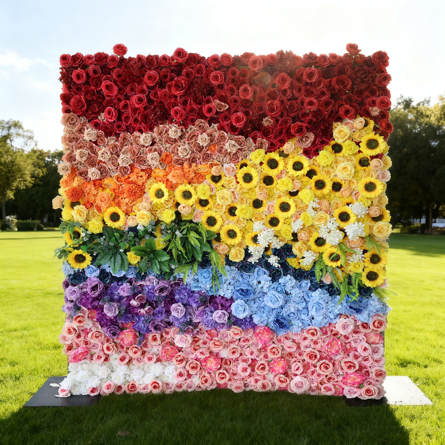 This image showcases a vibrant floral wall by Valar Flowers, set outdoors on a lush green lawn under a clear sky. It features a stunning arrangement of artificial flowers in a rainbow of colors—deep reds, soft creams, bright yellows with sunflowers, various blues, purples, and pinks—interspersed with lush green foliage. The layered, colorful design creates a lively and eye-catching display, perfect for adding a touch of natural beauty and charm to events or outdoor spaces.