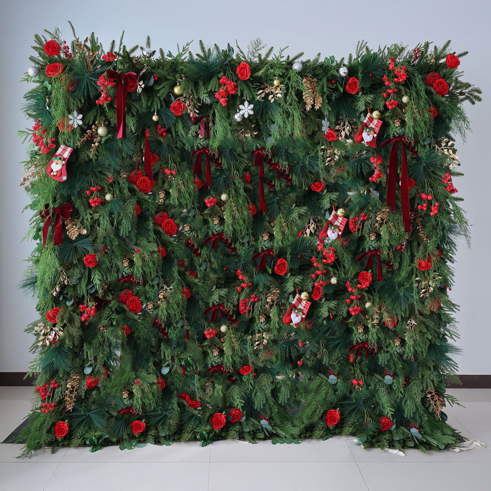 This image displays a large, festive Christmas - themed floral wall. It is richly decorated with lush green foliage, vibrant red roses, and various Christmas ornaments such as small stockings, snowflakes, and baubles. Deep red ribbons are also incorporated, adding to the holiday charm. Set against a plain light - colored wall and a tiled floor, the wall exudes a warm and joyful Christmas atmosphere, perfect for holiday decorations.