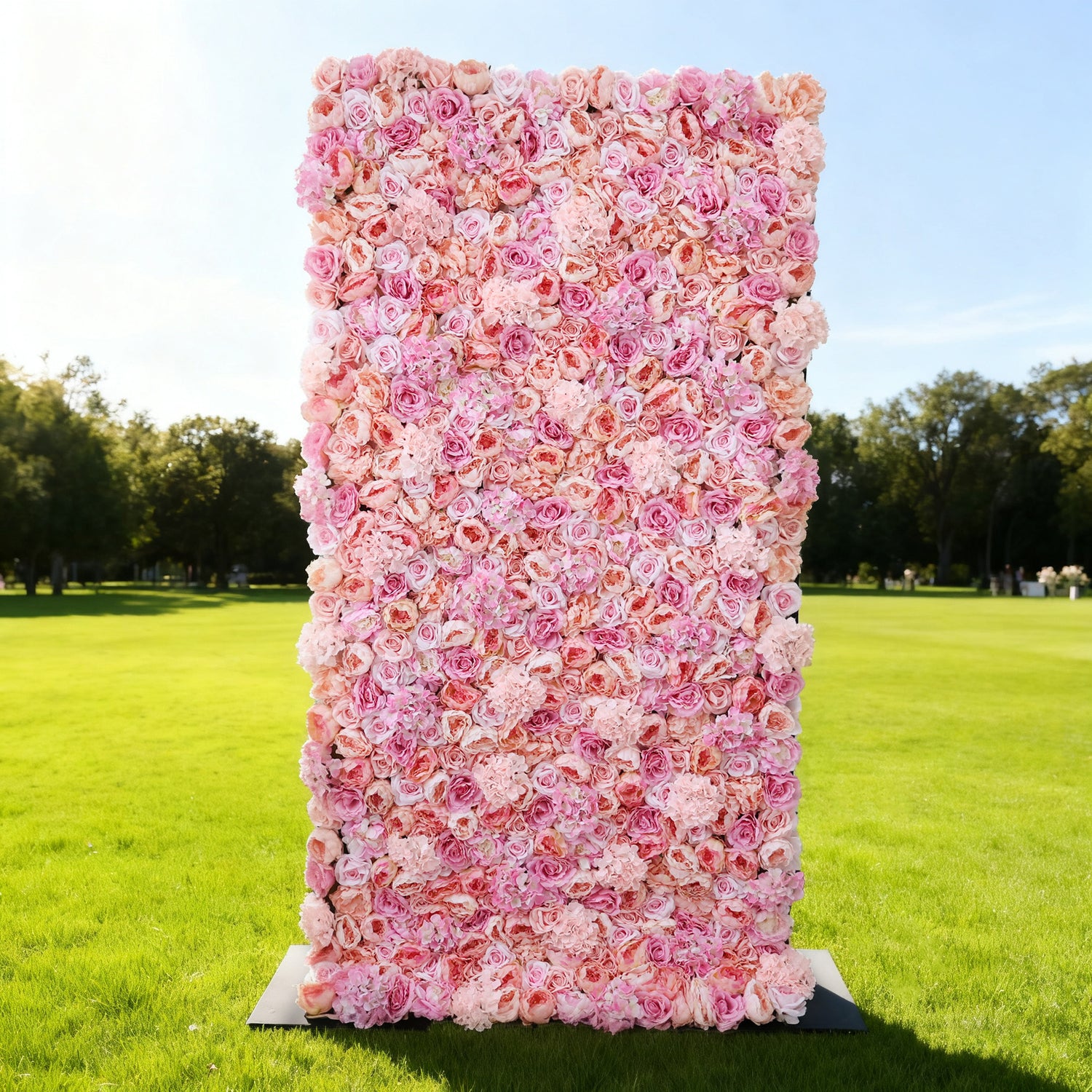 This image showcases a stunning pink floral backdrop set outdoors on a vibrant lawn. It features a dense arrangement of artificial flowers in various shades of pink, creating a lush, textured display. Bathed in sunlight with a scenic park in the background, it exudes elegance, perfect for adding a luxurious, nature-inspired element to outdoor events.