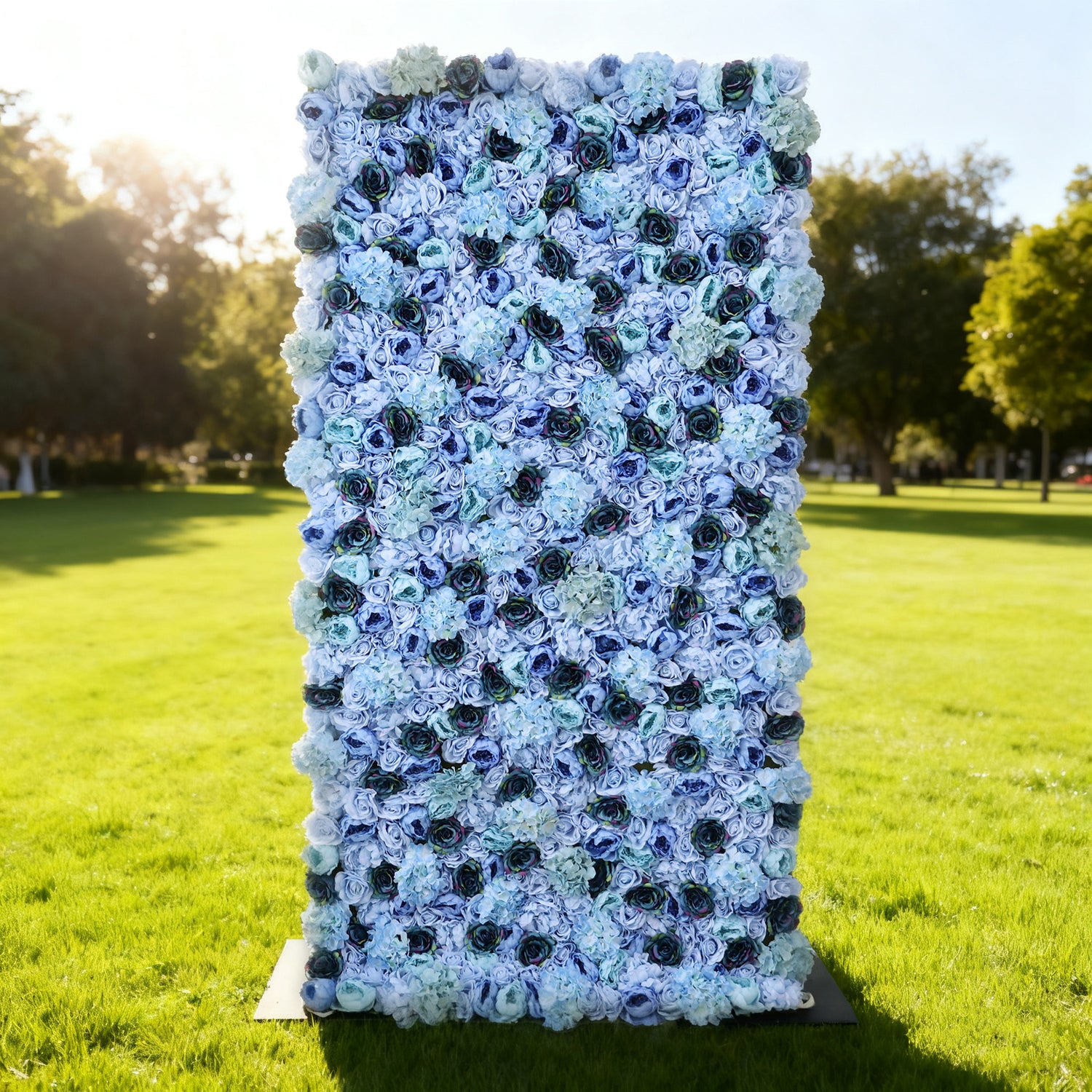 This image showcases a stunning floral backdrop by ValarFlowers, set outdoors on a vibrant lawn. It features a dense arrangement of artificial flowers in various shades of blue—from light sky blue to deep navy—including roses and hydrangeas, creating a lush, textured, and visually captivating display. Bathed in sunlight with a scenic park in the background, this piece exudes elegance and sophistication, perfect for adding a luxurious, nature-inspired element to outdoor events.