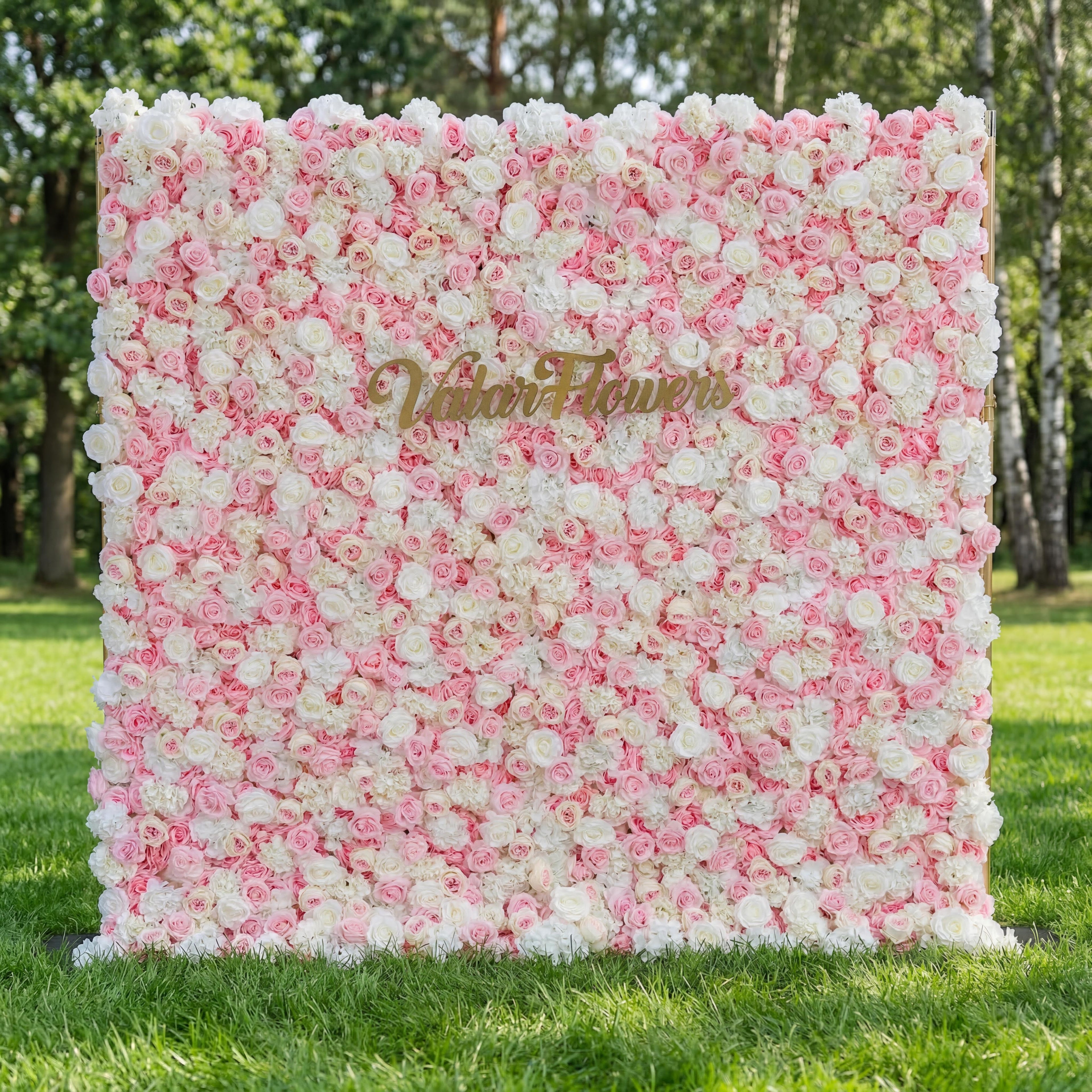 A stunning Valar Flowers floral wall on lush green grass features a dreamy gradient from crisp white at the top to soft blush and deep pink roses. Adorned with a gold "Valar Flowers" script sign, it creates an elegant, romantic backdrop perfect for weddings, bridal showers, and upscale photo sessions