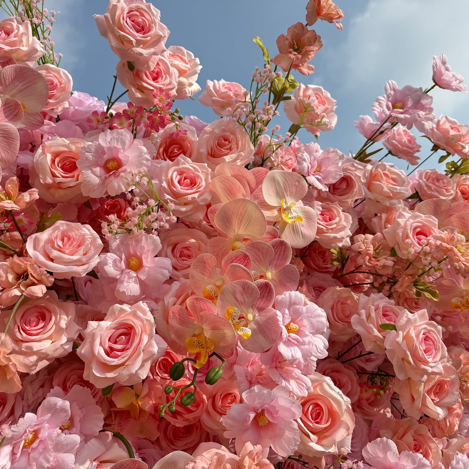 This photo showcases a lush and abundant floral arrangement filled with soft pink roses, delicate pink orchids, and small pink blossoms. The pastel - colored flowers create a gentle and dreamy atmosphere, set against a backdrop of a partly cloudy blue sky. The dense cluster of blooms exudes a sense of romance and elegance, perfect for events like weddings or high - end garden parties.
