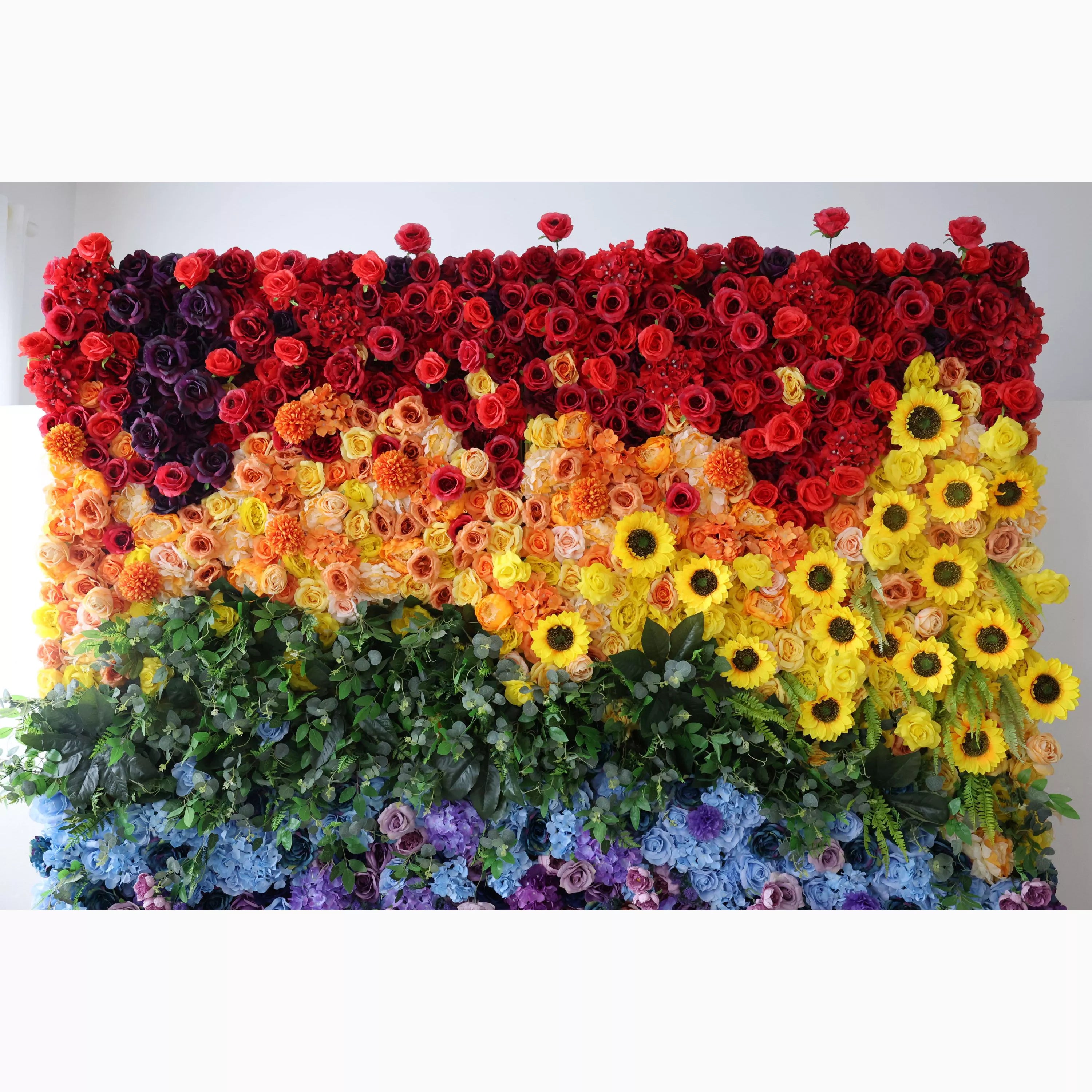 This vibrant indoor floral wall features a bold, sunset-to-rainbow gradient, transitioning from deep reds and rich purples at the top, through warm peaches and sunny yellow sunflowers, to lush green foliage and cool blue and purple blooms at the bottom. Its dense, textured arrangement of artificial flowers exudes joyful, vibrant energy, making it a lively focal point for pride events, festivals, or colorful celebrations against a minimalist white backdrop.