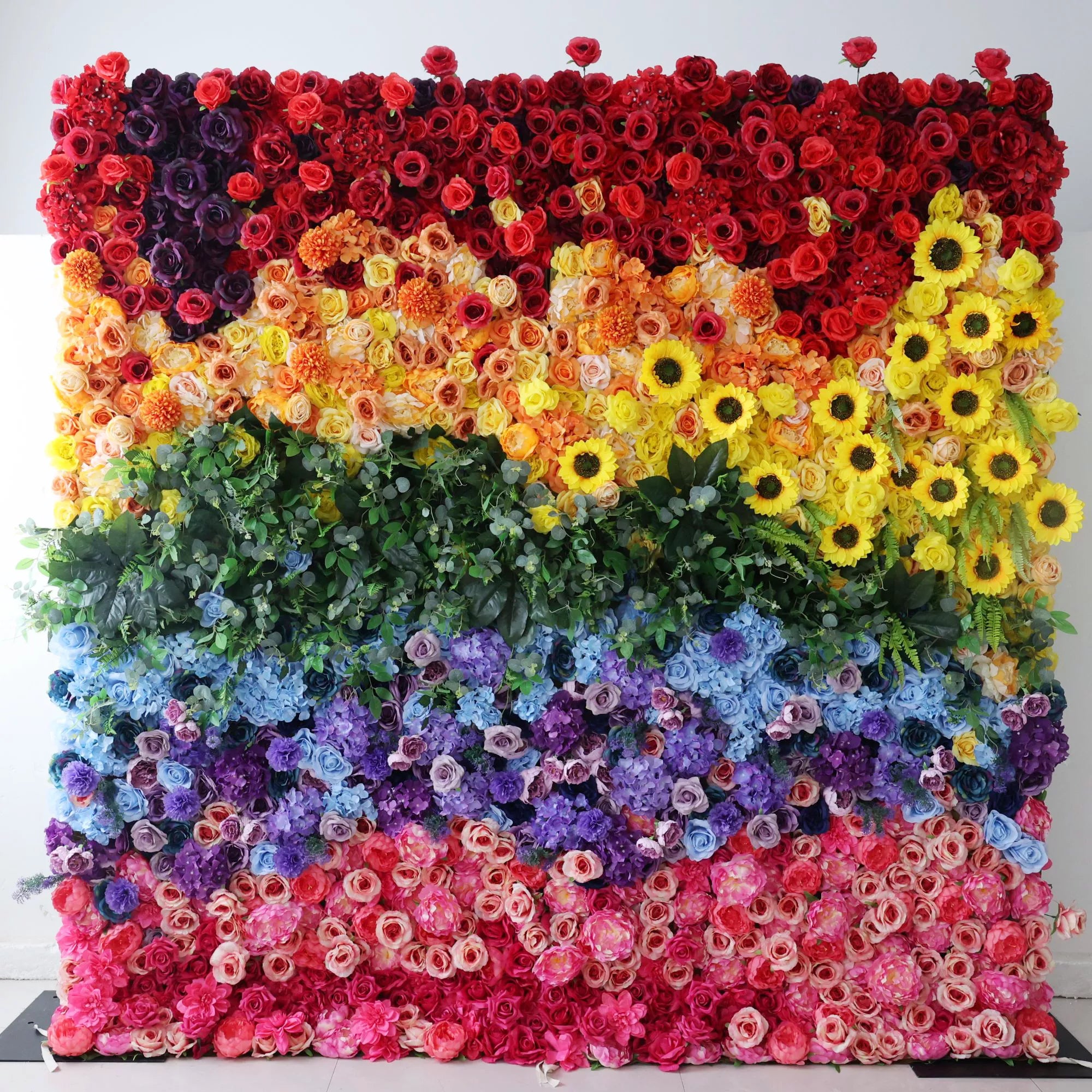 This vibrant floral wall features a bold, rainbow-inspired gradient, transitioning from deep reds and rich purples at the top, through warm peaches and sunny yellow sunflowers, to lush green foliage, cool blue and purple blooms, and soft pink and white roses at the bottom. Its dense, textured arrangement of artificial flowers exudes joyful, vibrant energy, making it a lively focal point for pride events, festivals, or colorful celebrations.