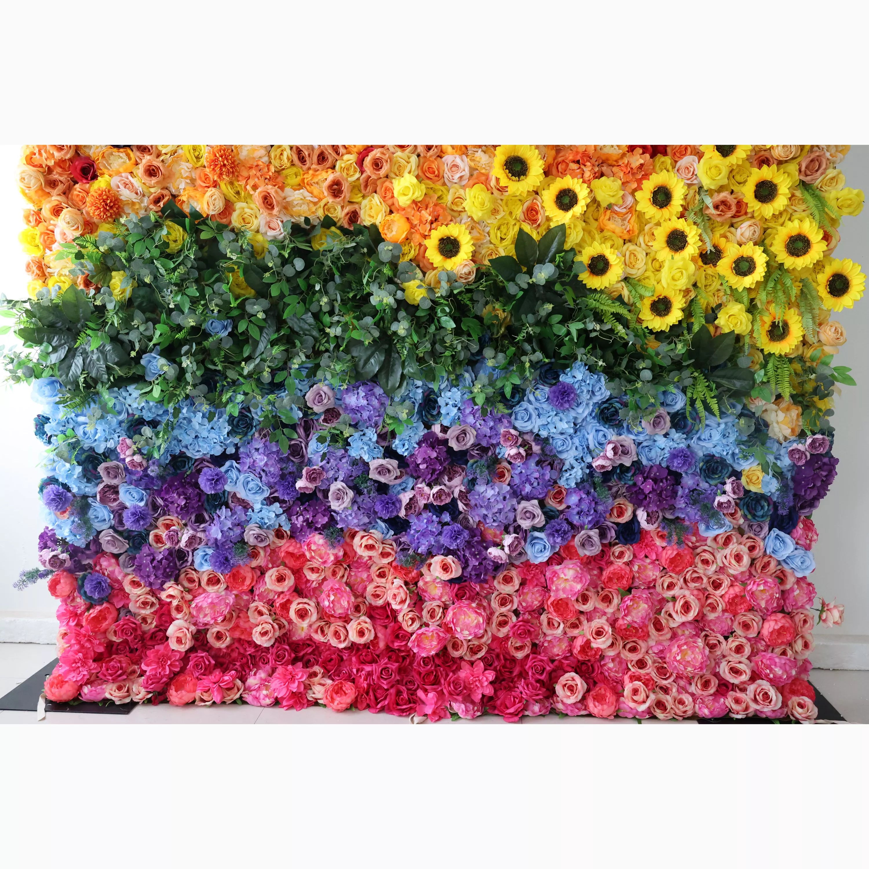 This vibrant floral wall features a dynamic, sunburst-style gradient, transitioning from warm peach and sunny yellow sunflowers at the top, through lush green foliage, to cool blue and purple blooms, and finishing with soft pink and deep red roses at the bottom. Its dense, textured arrangement of artificial flowers exudes joyful, energetic warmth, making it a lively focal point for pride events, festivals, or colorful celebrations.