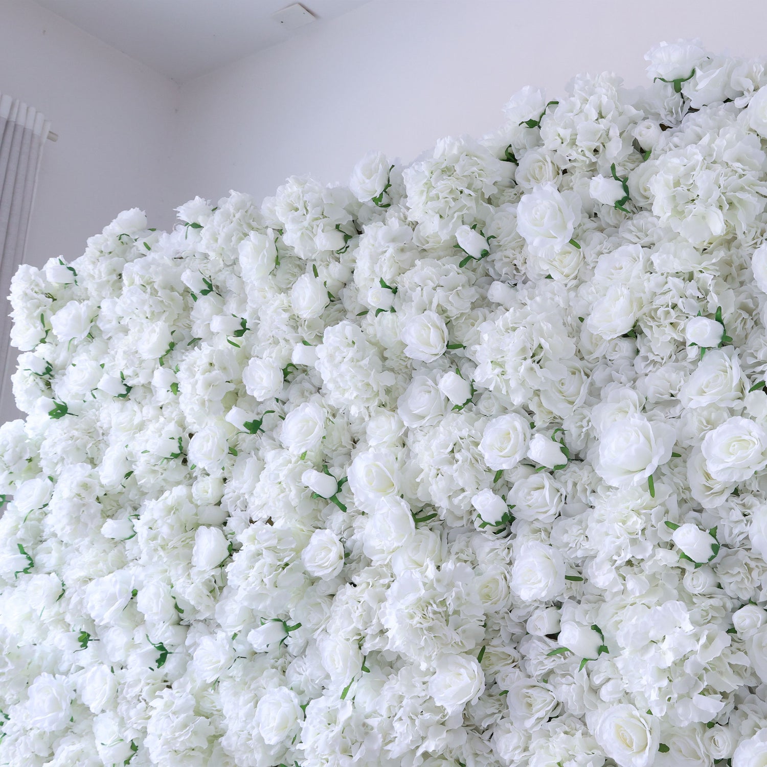 This image showcases a dense arrangement of white artificial flowers, creating a lush, voluminous floral display. The flowers, including roses and other blooms, are tightly packed, giving a sense of opulence and purity. Set against a plain white wall, the scene exudes an elegant and serene atmosphere, perfect for decorative purposes in events or as a striking interior backdrop, evoking a feeling of sophistication and natural beauty.