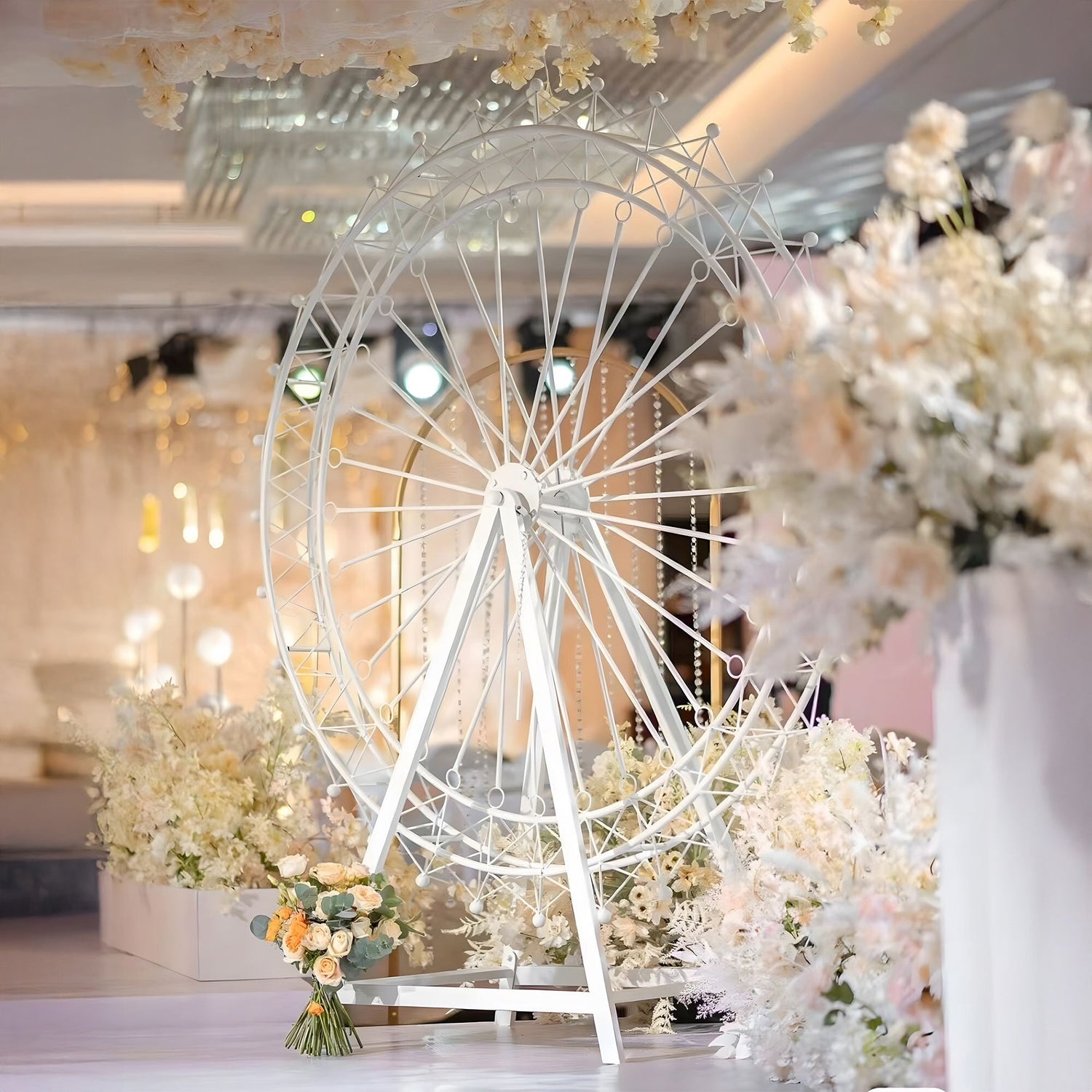 A large white Ferris wheel serves as a centerpiece at an elegant event, surrounded by soft - toned floral arrangements. Delicate beads dangle from its spokes, with warm lighting and crystal decor in the background, creating a romantic, dreamy atmosphere.