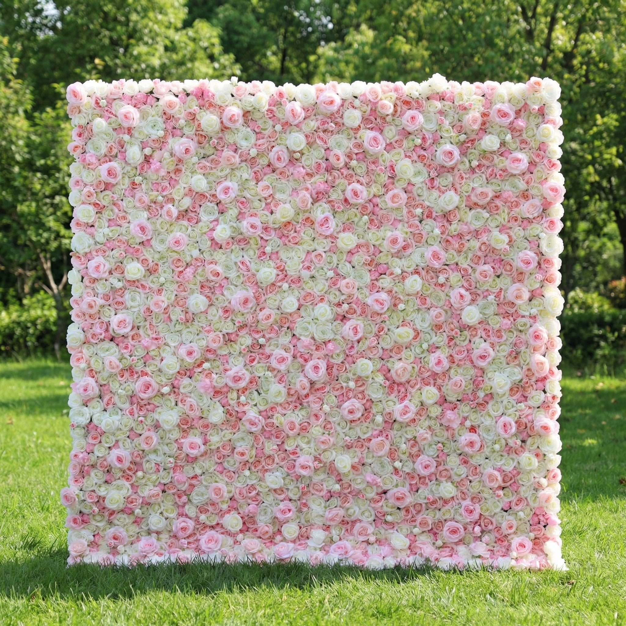 This is a large, rectangular artificial flower wall backdrop, densely packed with a mix of soft pink, white, and pale green roses and peonies. It stands upright on a lush green lawn, with a backdrop of leafy trees, creating a romantic, elegant setting ideal for weddings, photo shoots, or special events.