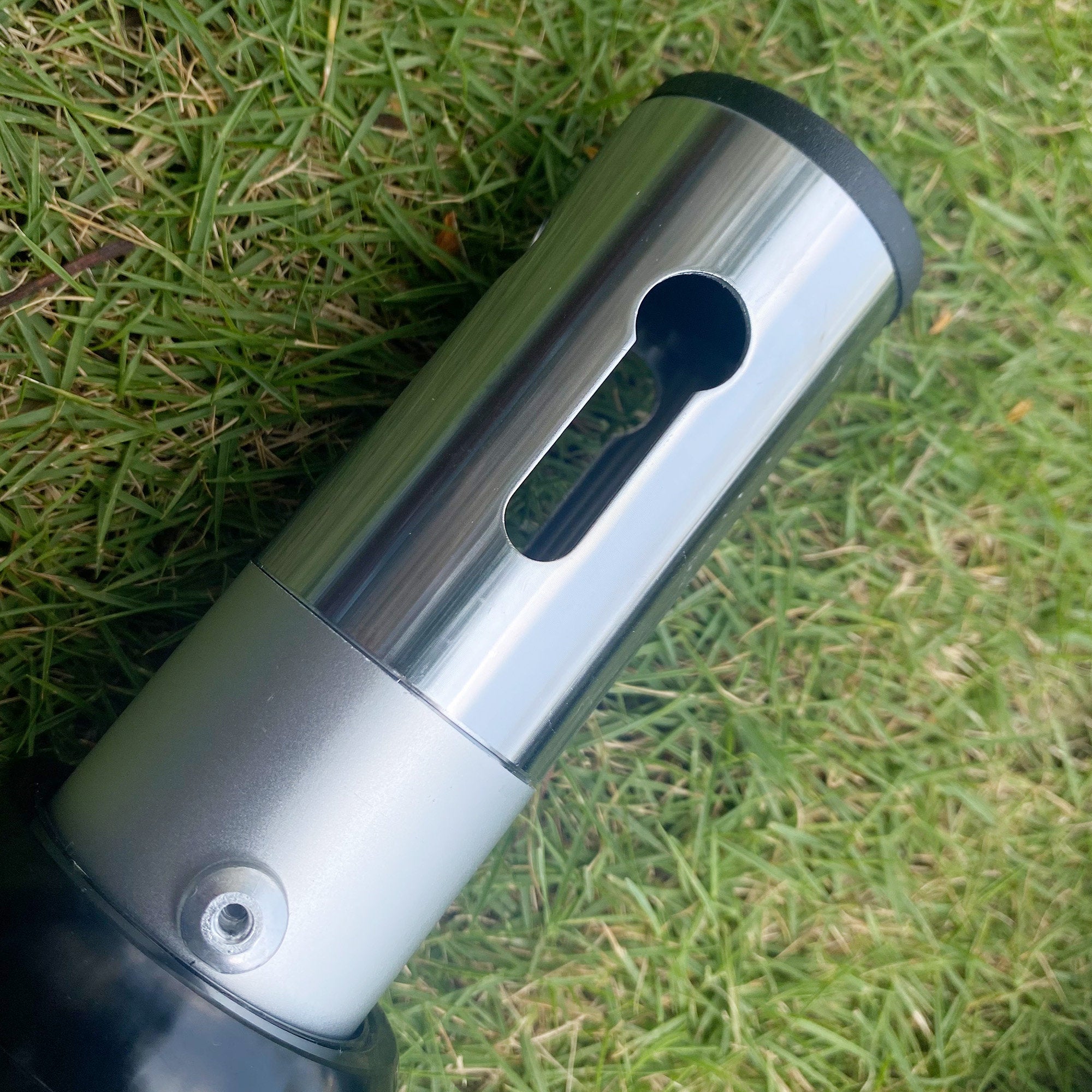 This image features a close - up of a metallic pole section on green grass. It has a rectangular cut - out in the upper part and a small circular screw or bolt near the bottom, likely part of an adjustable structure.