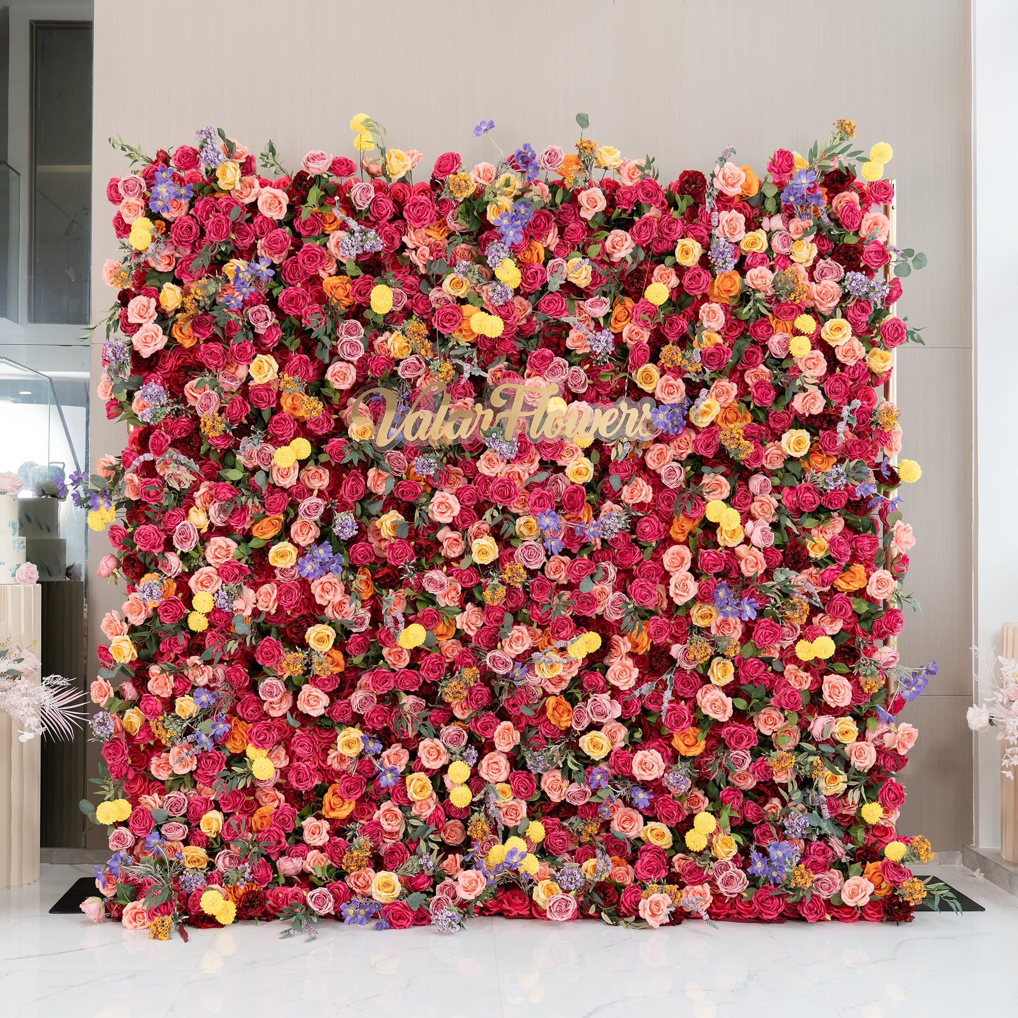 This image showcases a vibrant floral wall densely packed with artificial flowers. Predominant flowers are roses in shades of deep red, pink, and peach. Accents include yellow pom - pom flowers and small purple blooms. The word "ValarFlowers" in cursive script stands out. The style is lush and colorful, ideal for events or photo backdrops.