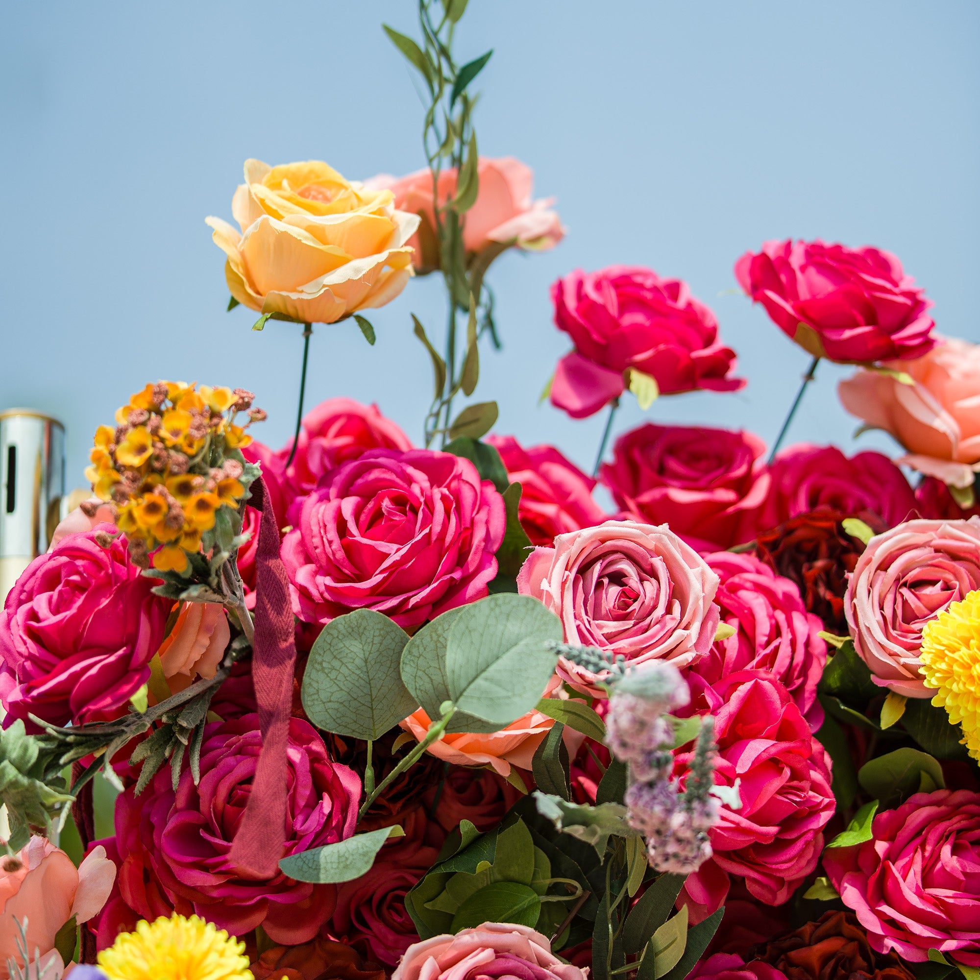 This vibrant floral arrangement features a lush mix of blooms. Deep pink roses take center stage, surrounded by soft peach - toned roses and sunny yellow flowers. Delicate yellow clusters and purple sprigs add texture, while eucalyptus leaves provide a fresh green contrast. The bright sky backdrop enhances the vivid colors, creating a lively, eye - catching display perfect for celebrations or decorative focal points.