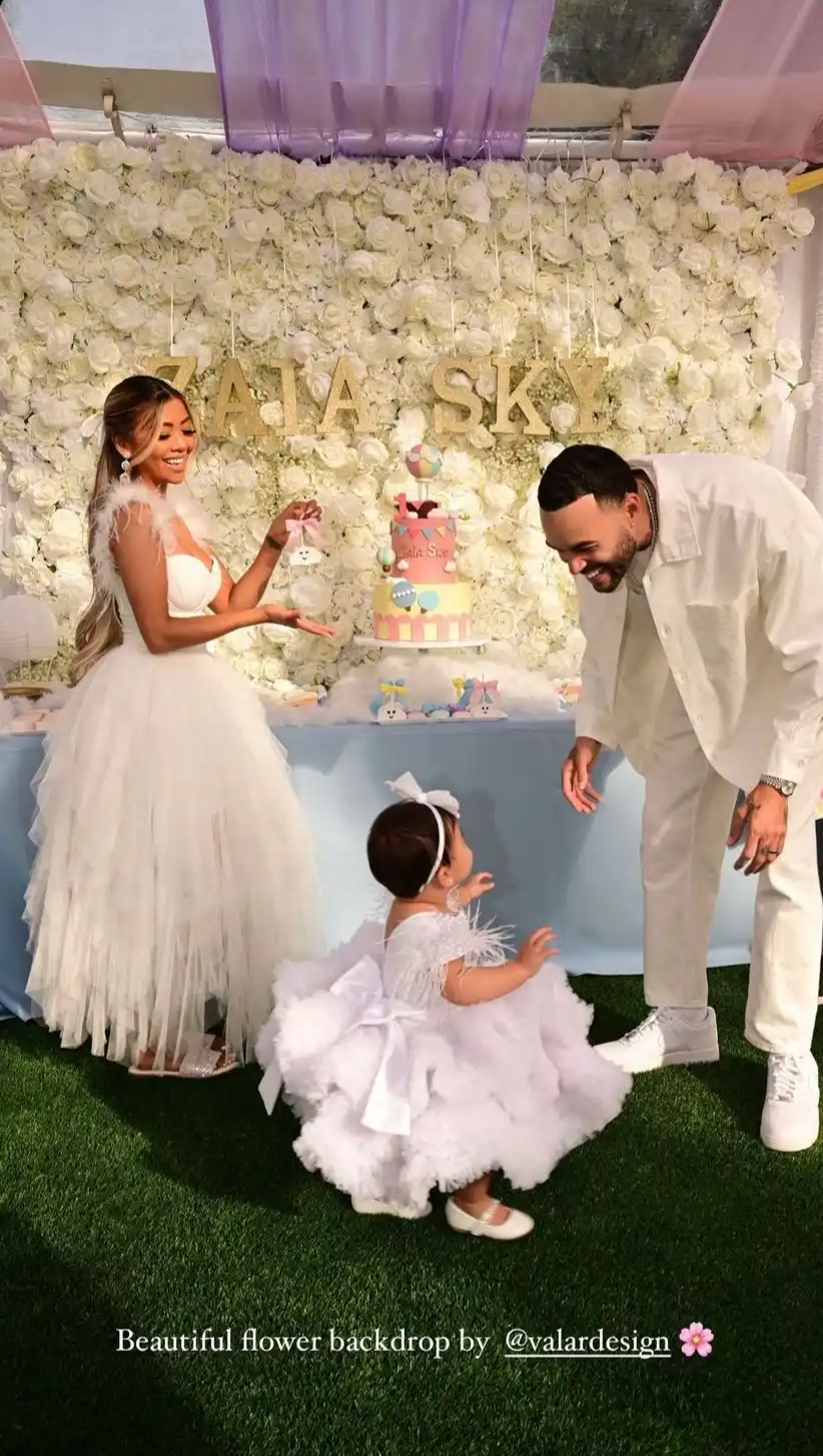 A joyful outdoor scene with a family in white - themed outfits. A young girl in a frilly dress, a woman in a voluminous gown, and a man in a white suit are in front of a white floral backdrop saying "ZAIA SKY". There's a colorful cake, suggesting a celebratory event.