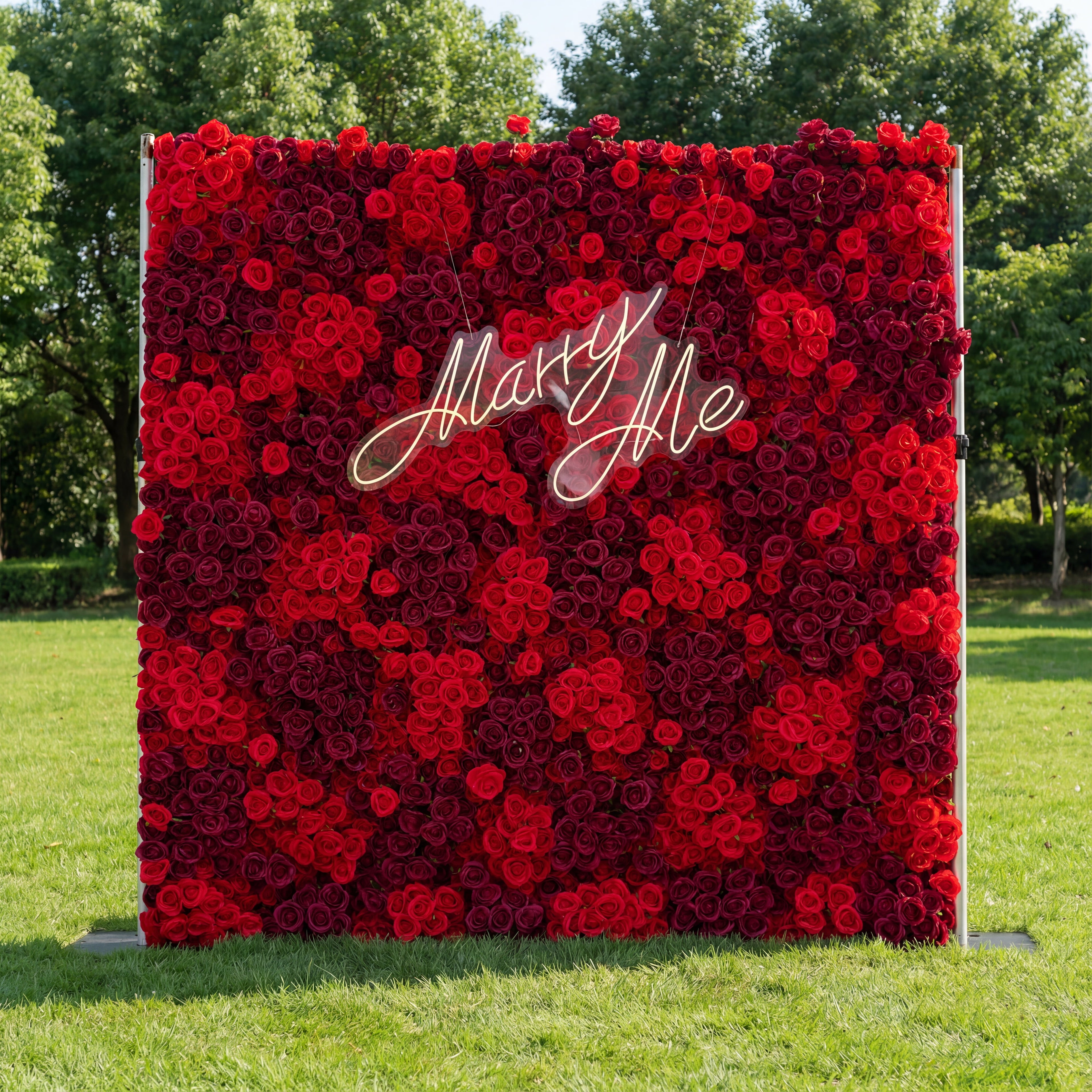 This striking backdrop (set on sunlit grass, tree-lined background) is dense with layered red roses—deep burgundy to bright crimson—creating a lush, romantic texture. A glowing "Marry Me" neon sign (cursive, light-lit) anchors the center, contrasting vividly with the rich red blooms. The bold, monochromatic palette evokes passion and elegance, making it ideal for proposal setups, romantic events, or wedding decor.